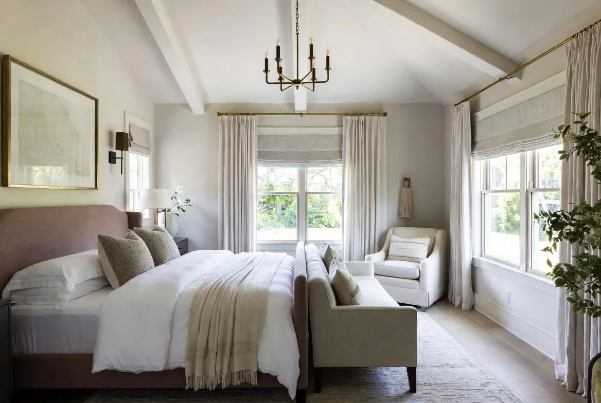Cuarto con cama grande con cabecera de tela en color rosado, dos ventanas con cortinas blancas, sillón de brazos en color blanco, sofá, luz natural, lámpara de mesa y lámpara de pared, decoraciones sencillas y ambiente luminoso.