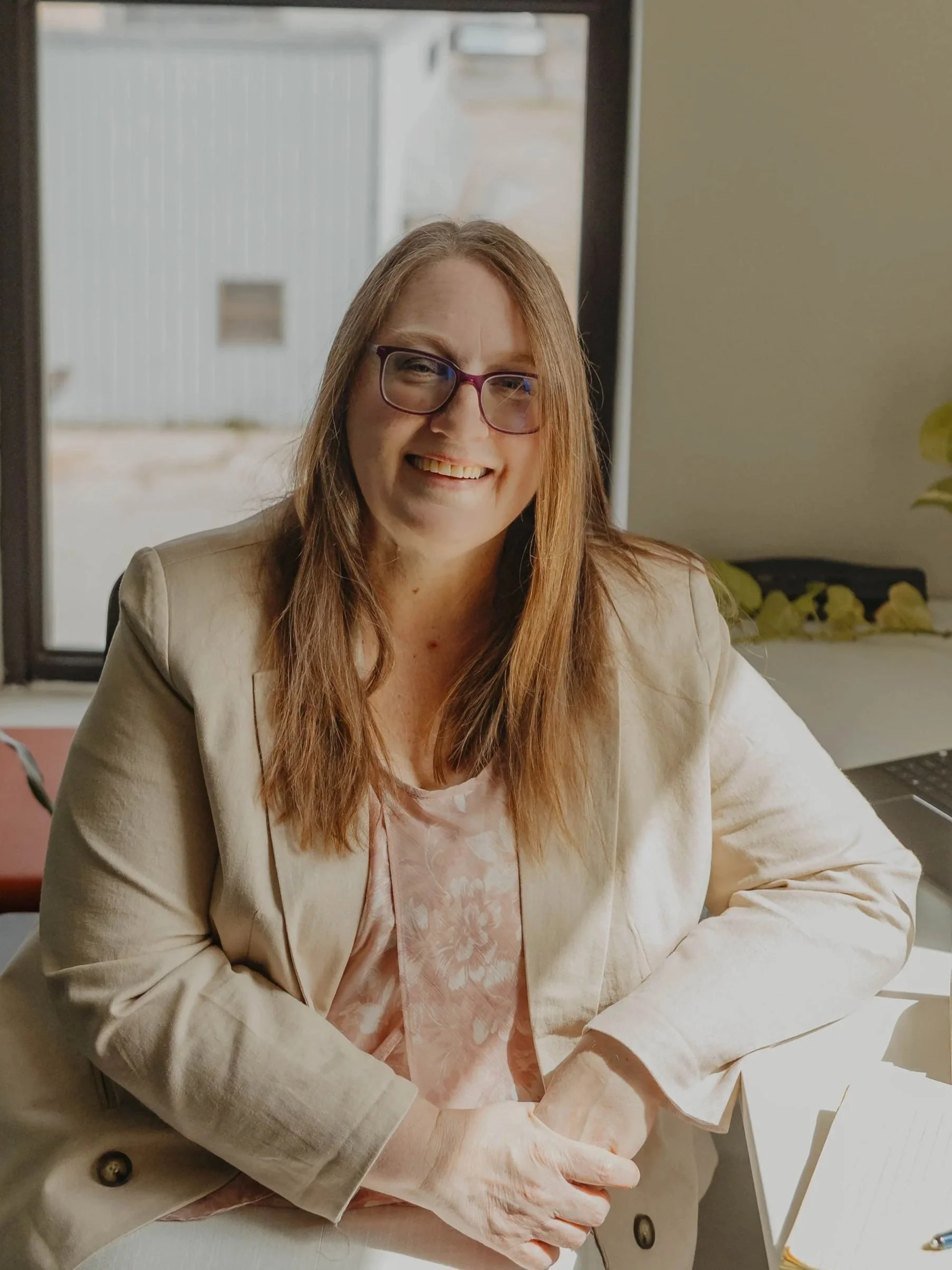 A woman with long red hair and glasses sitting at a desk near a window, smiling at the camera.