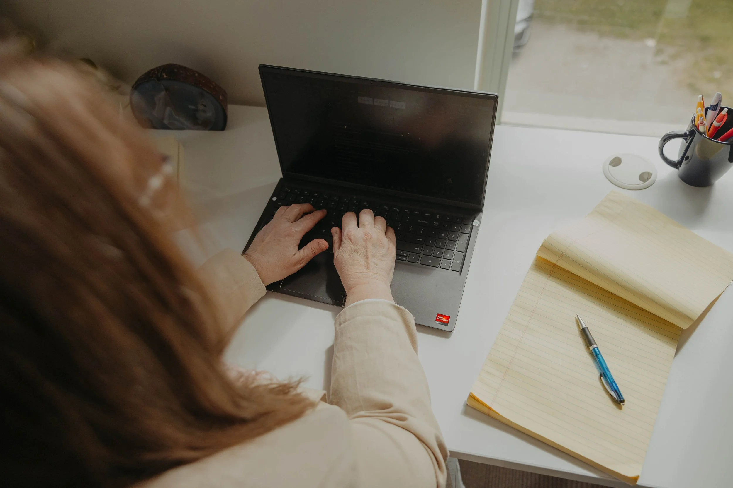 Person typing on a laptop computer at a white desk with a yellow notepad, pen, and a mug filled with colorful pens, near a window.
