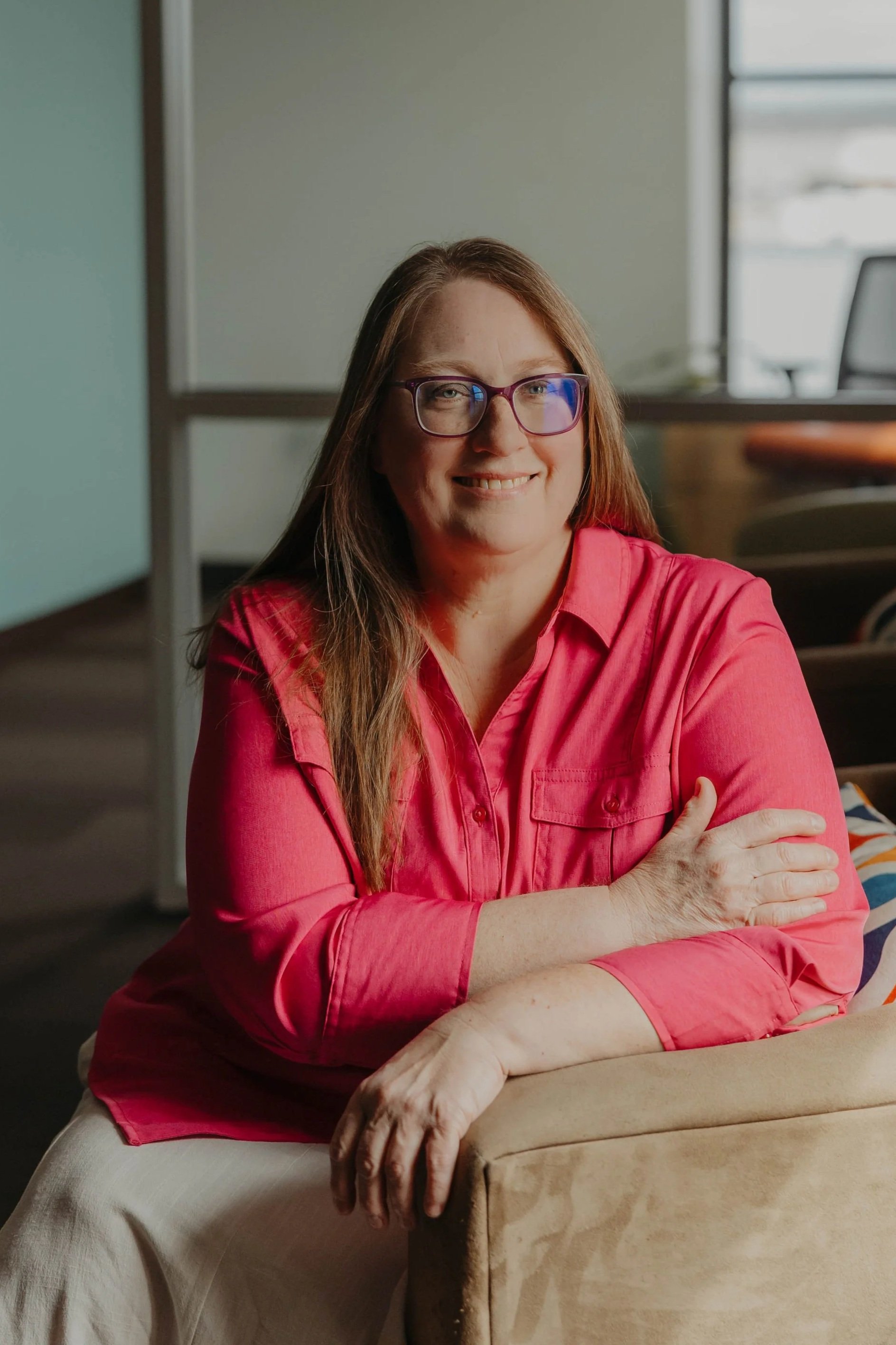 A woman with long brown hair, wearing glasses and a bright pink shirt, smiling with arms crossed, sitting in a room with a window and a soft background.
