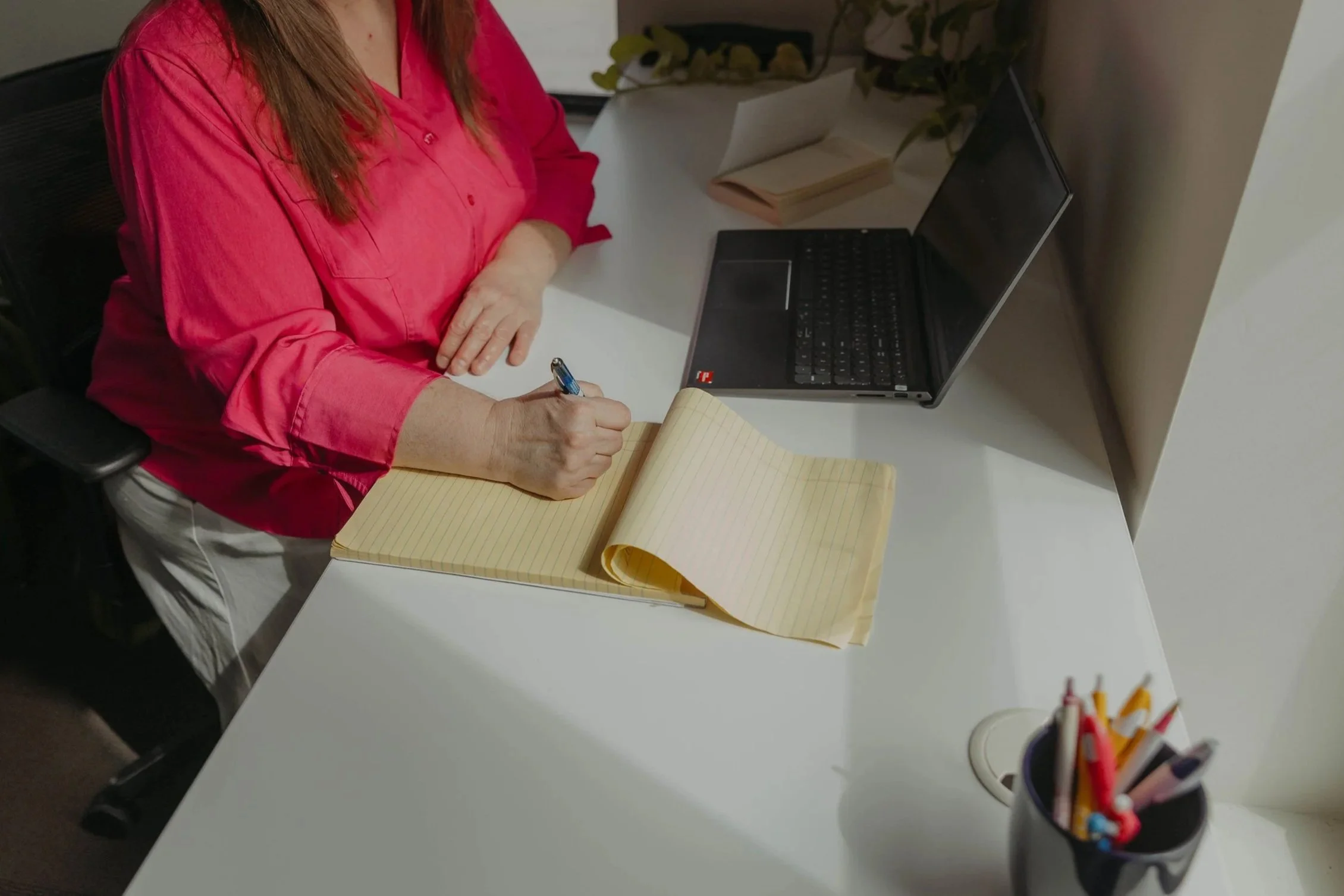 A person in a pink shirt is seated at a white desk, writing in a yellow notepad with a blue pen. There is an open laptop, a closed notebook, a pen holder with various pens and markers, and some books in the background.