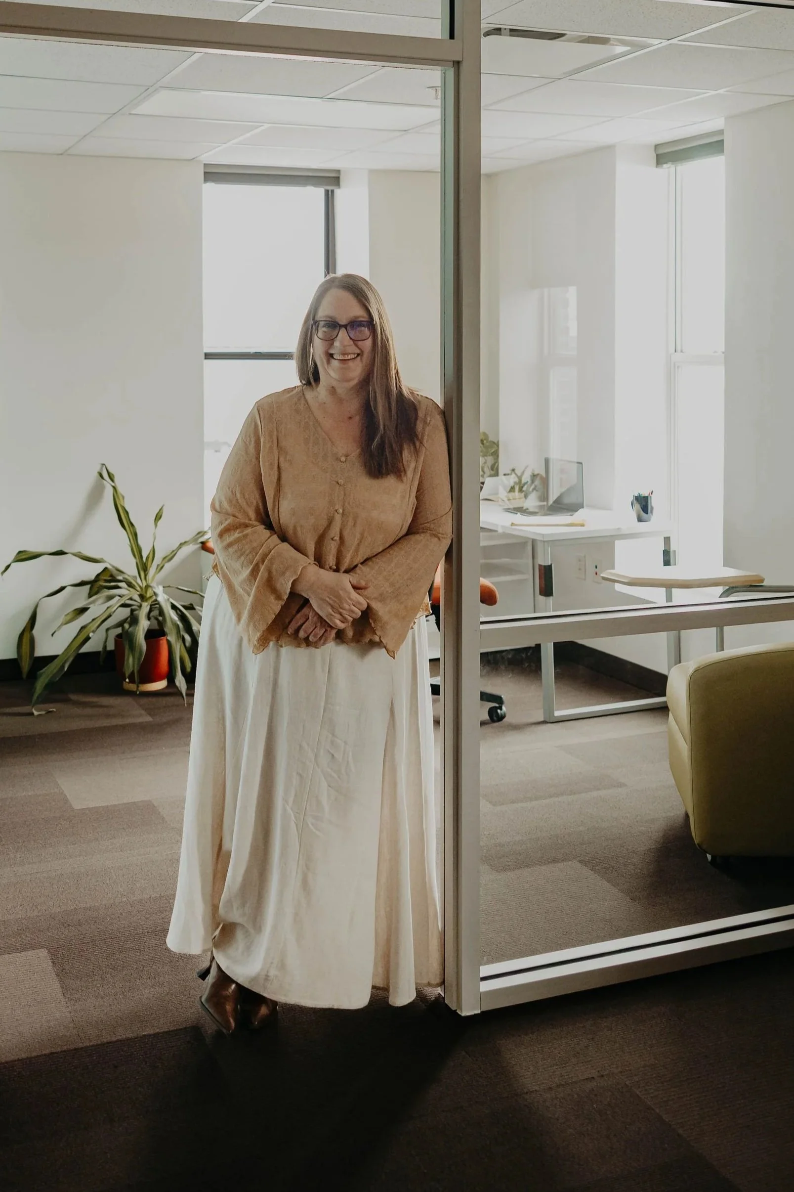 A woman with glasses and long brown hair standing in an office behind a glass partition wall, smiling at the camera. She wears a tan blouse and a long cream-colored skirt. The office has large windows, a desk with a computer, some plants, and office supplies.