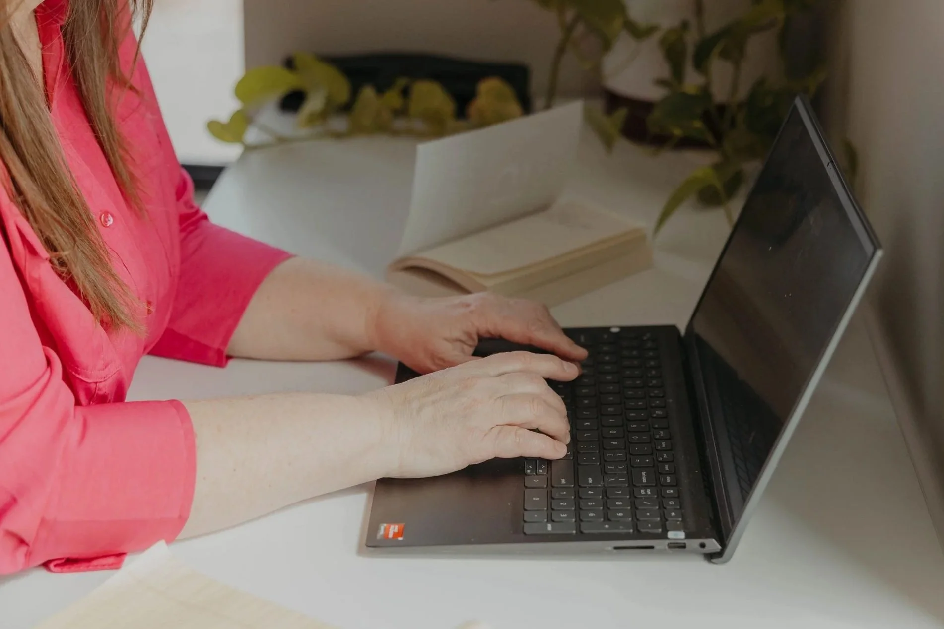 Close-up of elderly woman's hands typing on a black laptop keyboard, wearing a pink blouse. An open book and some potted plants are in the background.