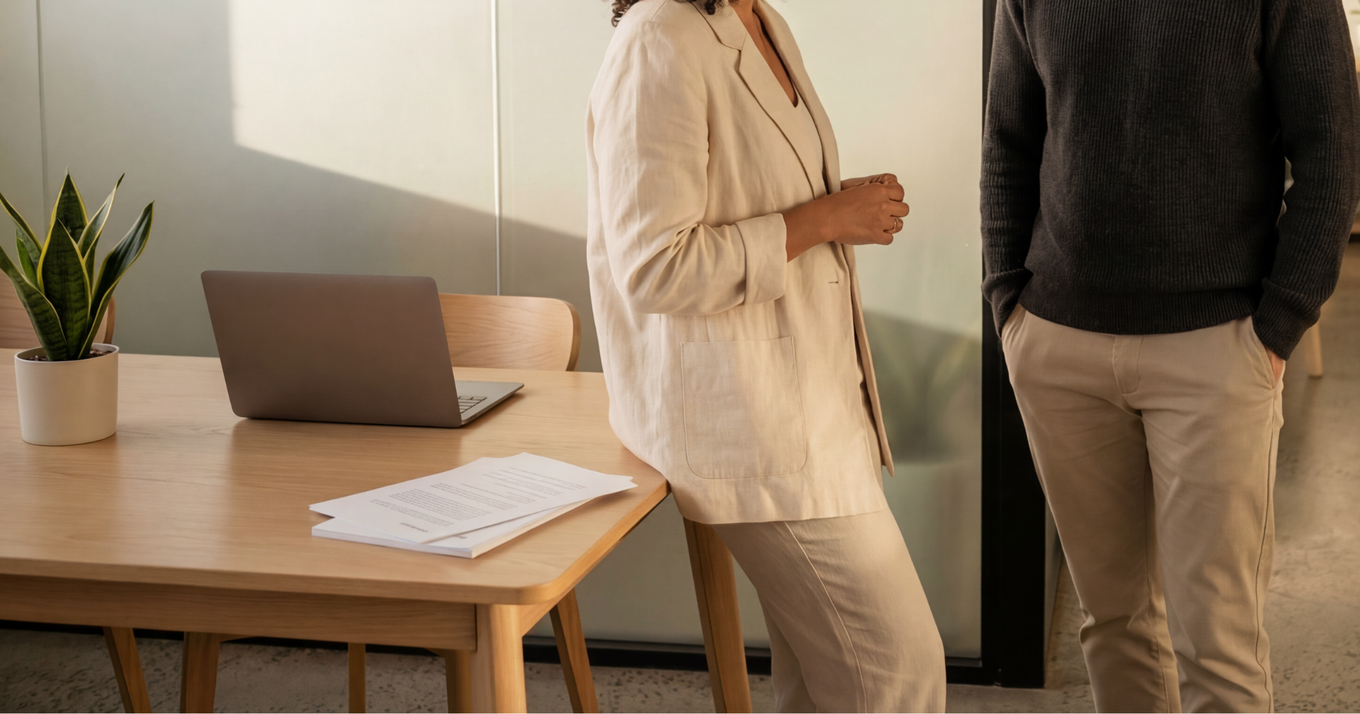Two people having a conversation in an office with a wooden table, an open laptop, and papers on it. There is a potted snake plant on the table and a glass partition in the background.