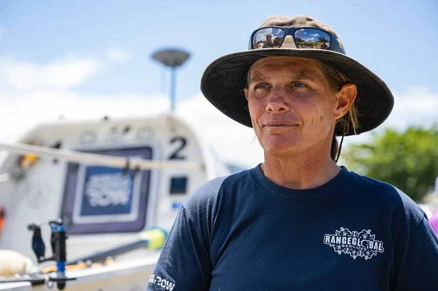 A woman wearing a wide-brimmed hat, sunglasses, and a navy blue t-shirt standing outdoors, with a boat and blue sky in the background.