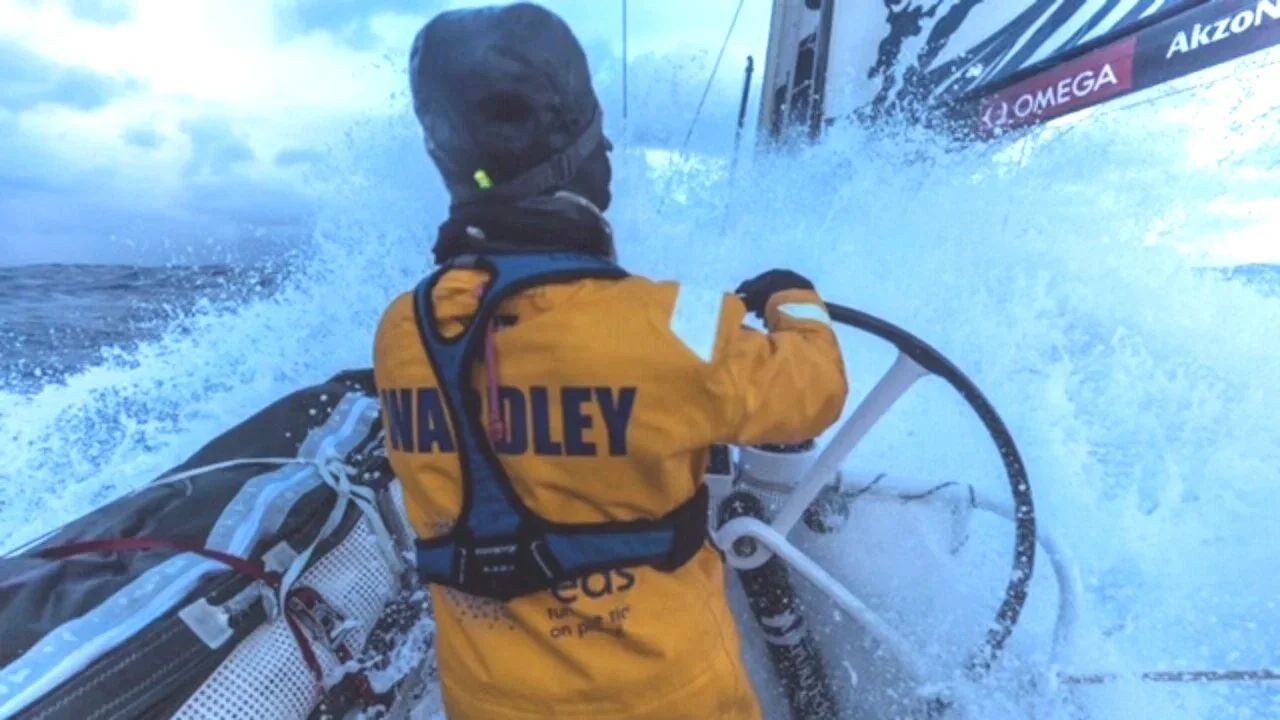 Liz Wardley steering an offshore racing yacht through rough seas in yellow waterproofs.
