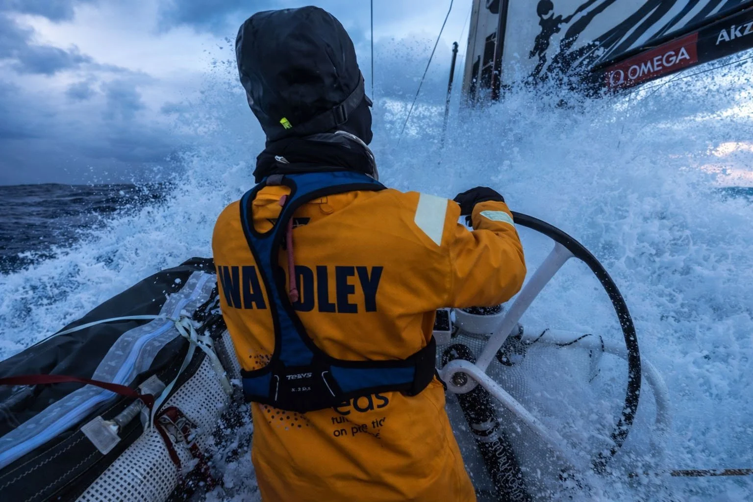 Liz Wardley steering an offshore racing yacht through rough seas in yellow waterproofs.
