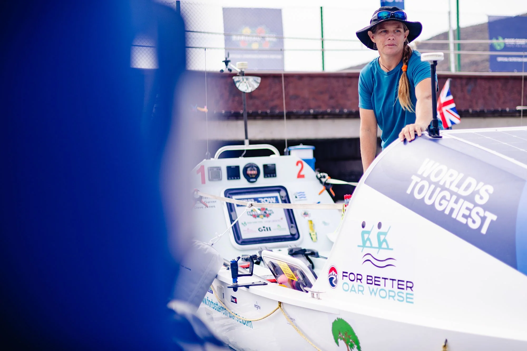 A woman wearing a blue shirt and hat working on a solar-powered boat during a race. The boat is white with various sponsor logos and a sign that reads "World's Toughest". The background shows a cityscape with mountains and a small Union Jack flag.