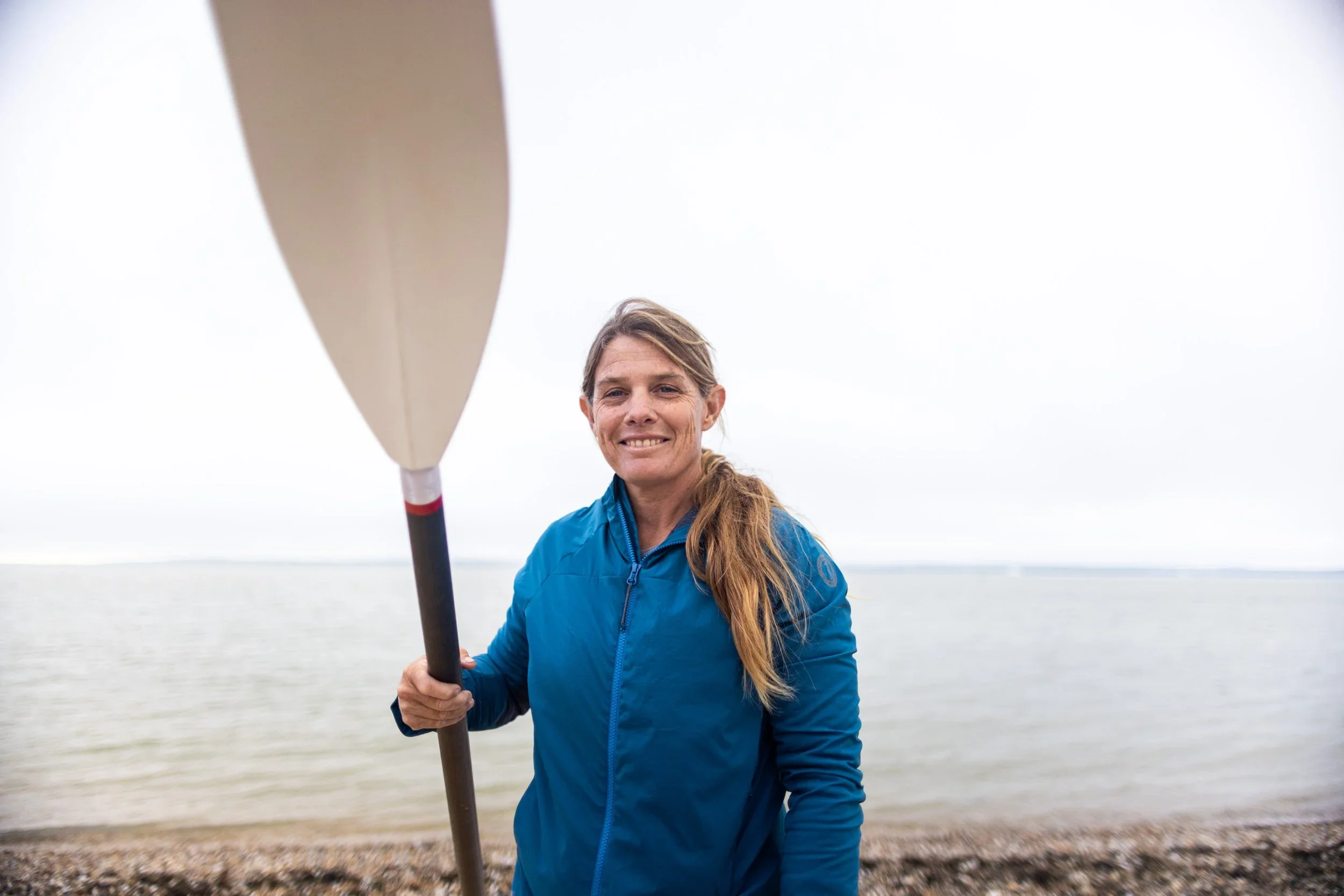 Woman with long hair in a ponytail wearing a blue jacket standing on a beach, holding an oar, with water and an overcast sky in the background.