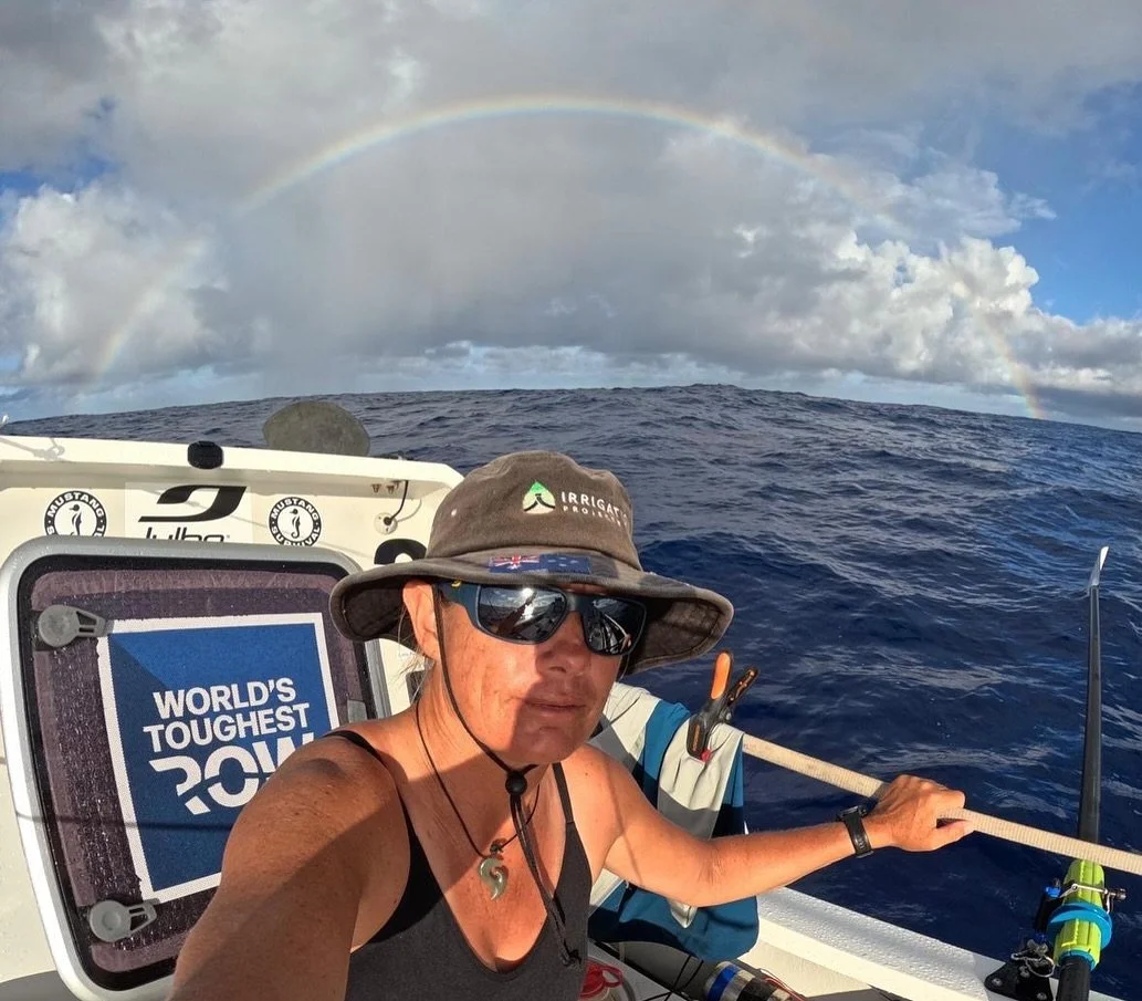 A person taking a selfie on a boat in the ocean, with a rainbow arching across the cloudy sky in the background.