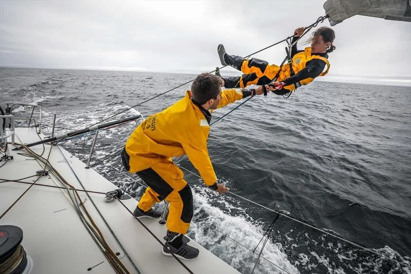 Two people in yellow waterproof suits rescue a person from a sailboat at sea, with one person holding the person in the air and the other securing the rescue line.