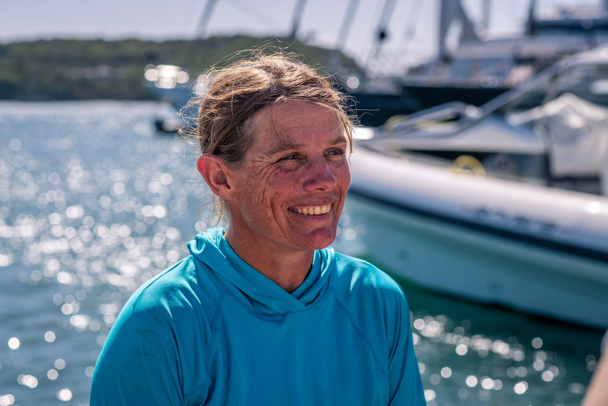A smiling woman with brown hair tied back, wearing a blue jacket, on a boat with a blurred background of water and other boats.