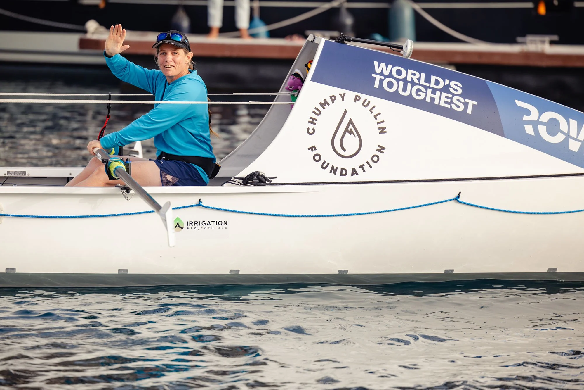 A woman in a blue jacket and sunglasses sitting on a sailboat, waving and smiling. The sailboat has a sign that reads 'World's Toughest' and a logo for the Chumpy Pullin Foundation.
