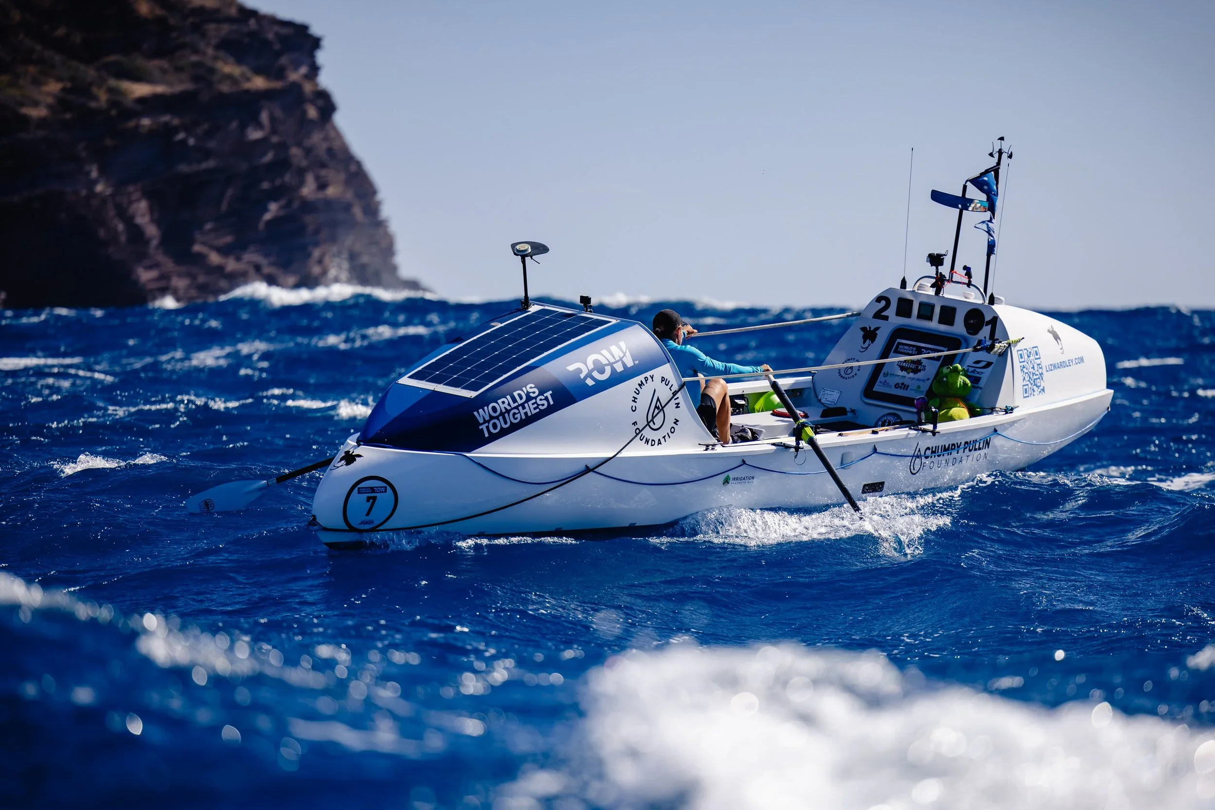 Liz Wardley rowing her solo ocean rowing boat near a rocky coastline during the World’s Toughest Row.