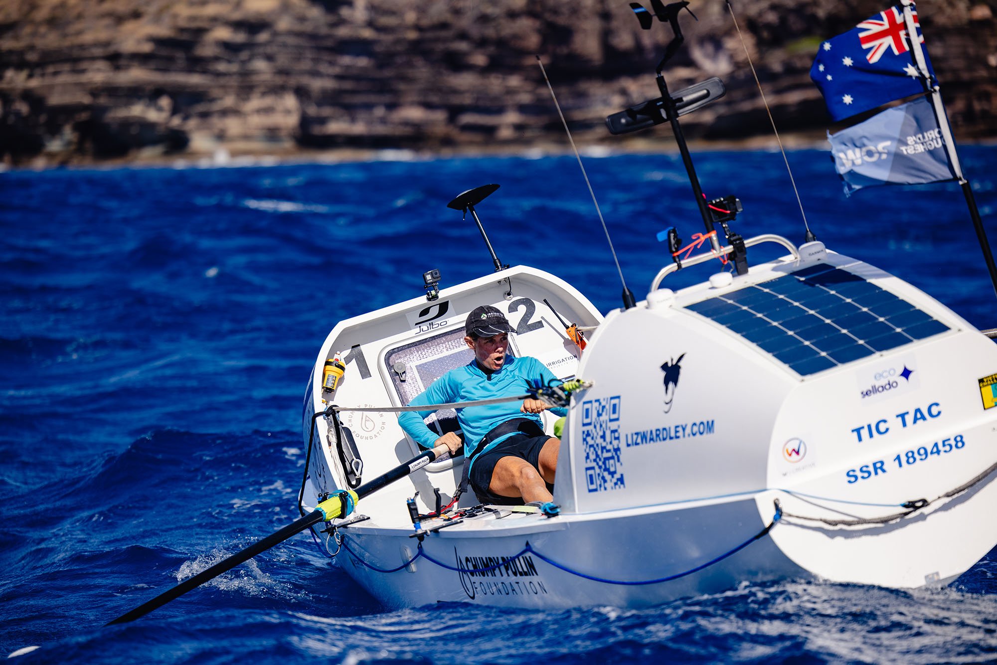 A person riding an electric boat with solar panels on it on choppy blue waters, with rocky coastline in the background, flying Australian and other flags.