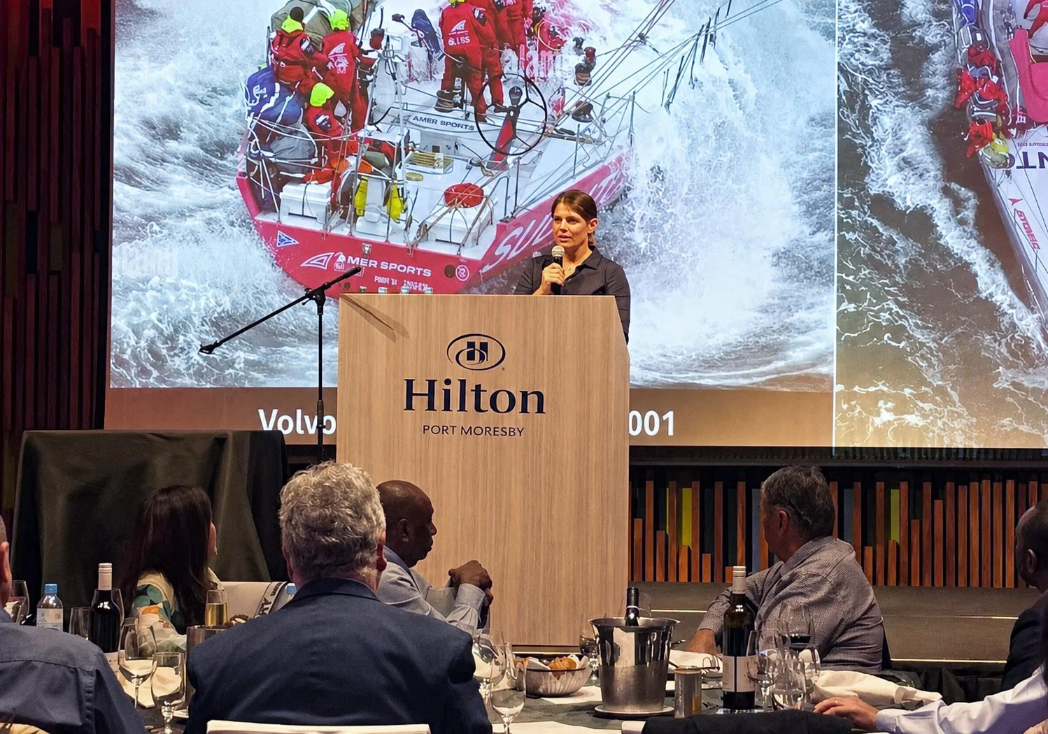 Liz Wardley speaking to an audience at Hilton Port Moresby, with an sailing boat displayed behind her.