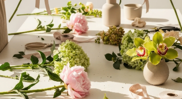 Arrangement of various flowers including pink peonies, green hydrangeas, orchids, and greenery on a white surface with decorative vases and natural light.