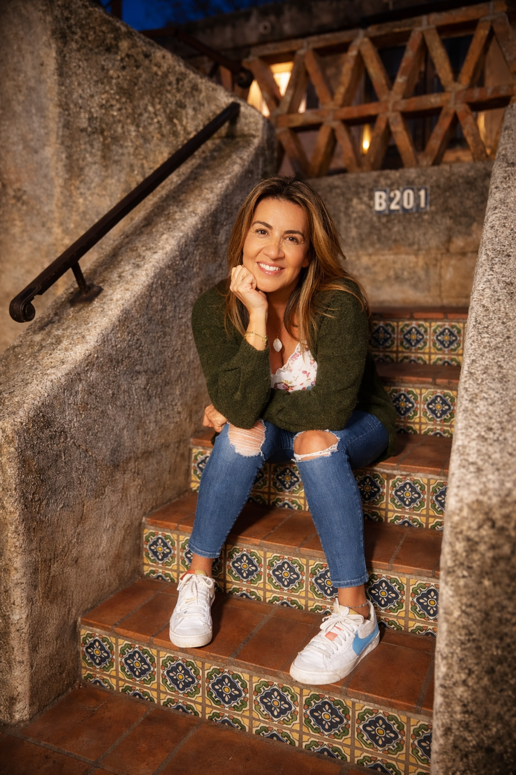 A woman smiling and crouching on colorful tiled stairs in a rustic setting, wearing white sneakers, ripped jeans, a floral top, and a green cardigan.