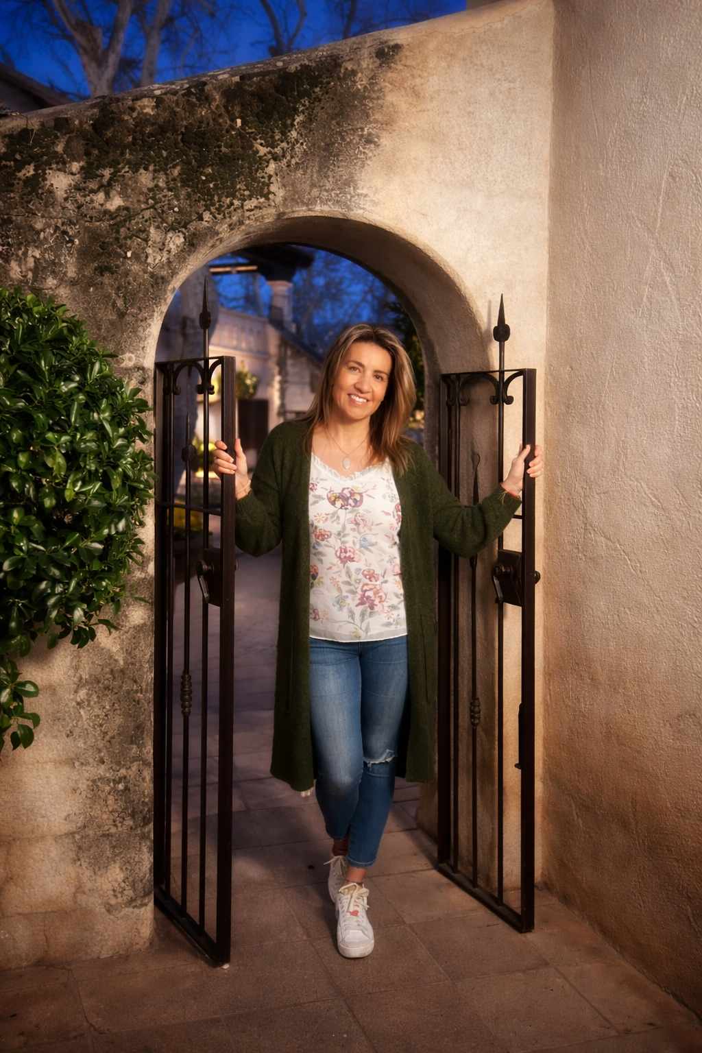 A woman with long brown hair, wearing a floral shirt, dark green cardigan, jeans, and white sneakers, standing in an open iron gate at dusk in a courtyard with stone and stucco walls, smiling and holding the gate open.