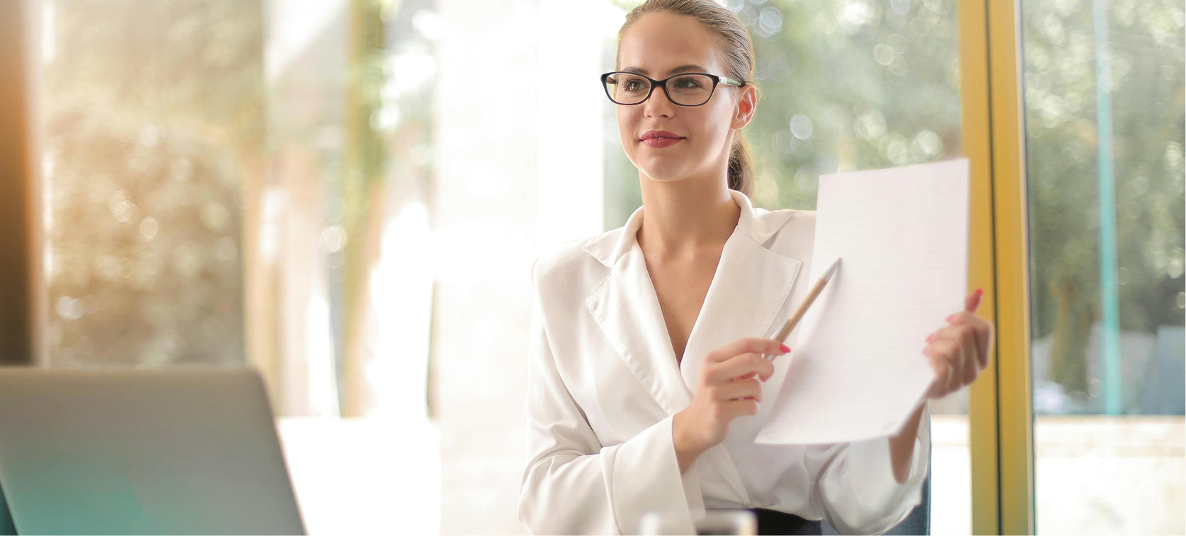 A woman wearing glasses, a white blazer, and pink lipstick holding a notepad and a pencil, sitting by a large window with a blurred outdoor background.