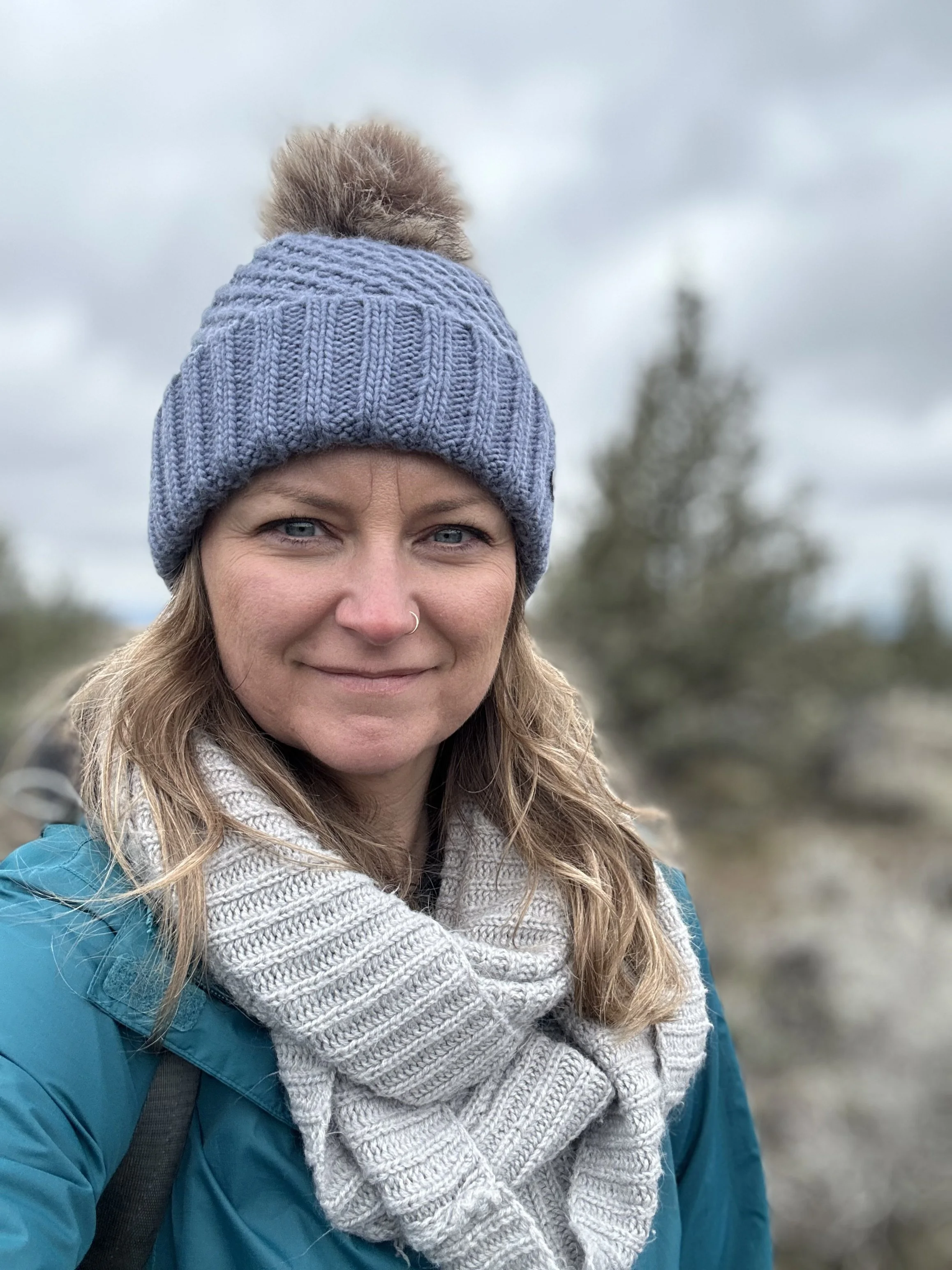 A woman with blonde hair wearing a gray knit beanie with a pom-pom, a gray scarf, and a blue jacket outdoors on a cloudy day.