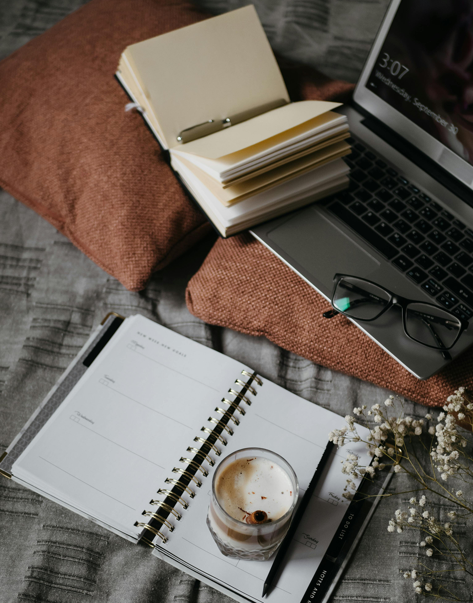 A workspace setup with a laptop, eyeglasses, an open planner, a glass of creamy drink, and a notepad with white flowers nearby on a textured gray and rust-colored fabric surface.