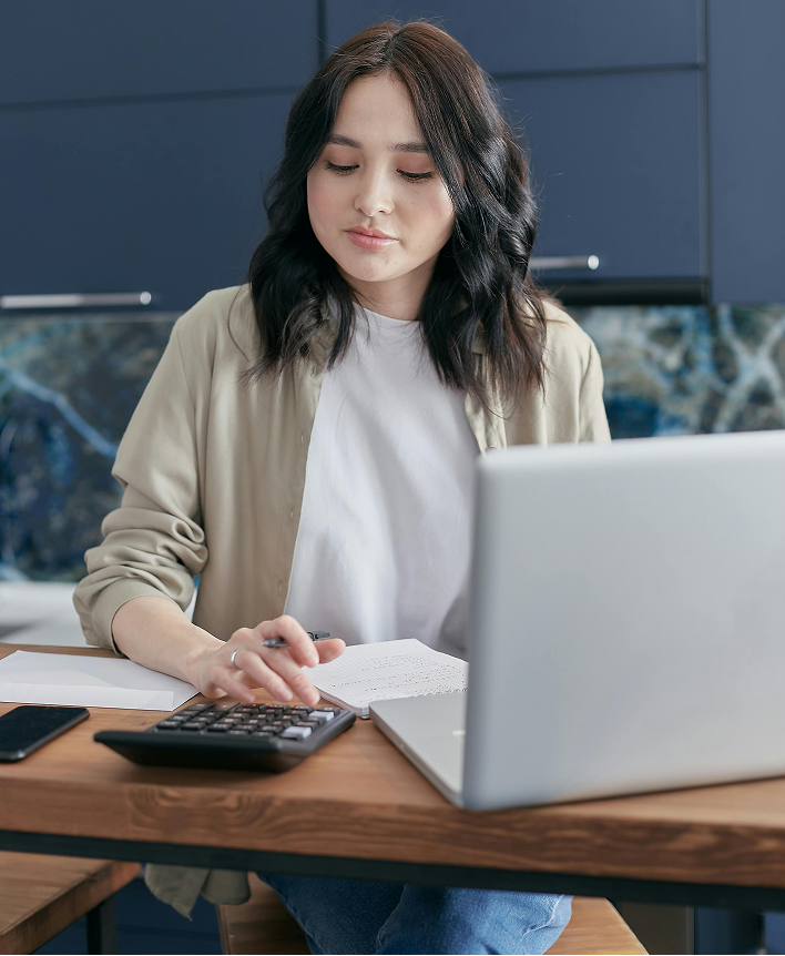 A young woman working at a desk with a laptop, calculator, and notebook in a modern kitchen or office space.