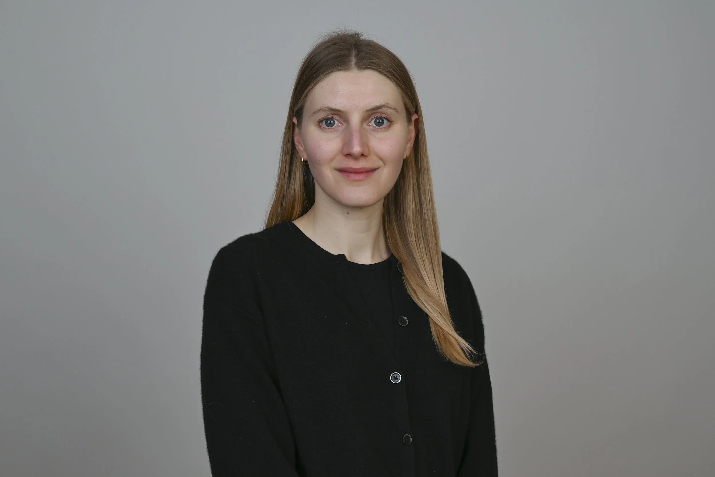 Portrait d'une femme aux yeux bleus, cheveux longs et blonds, portant un haut noir, contre un fond gris.