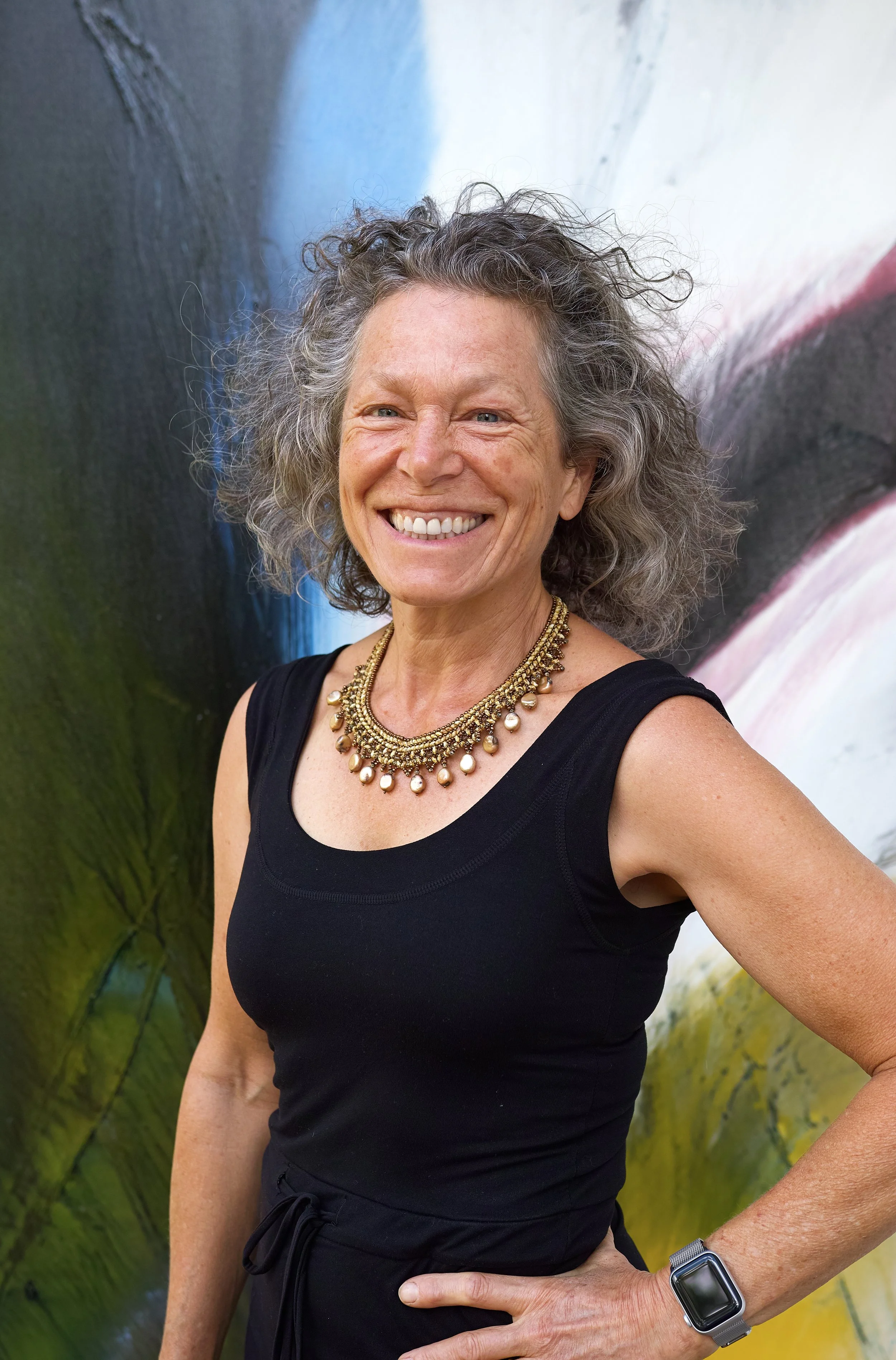 A smiling woman with curly gray hair, wearing a black sleeveless top, a layered pearl and gold necklace, and a silver smartwatch, standing in front of an abstract, colorful background.
