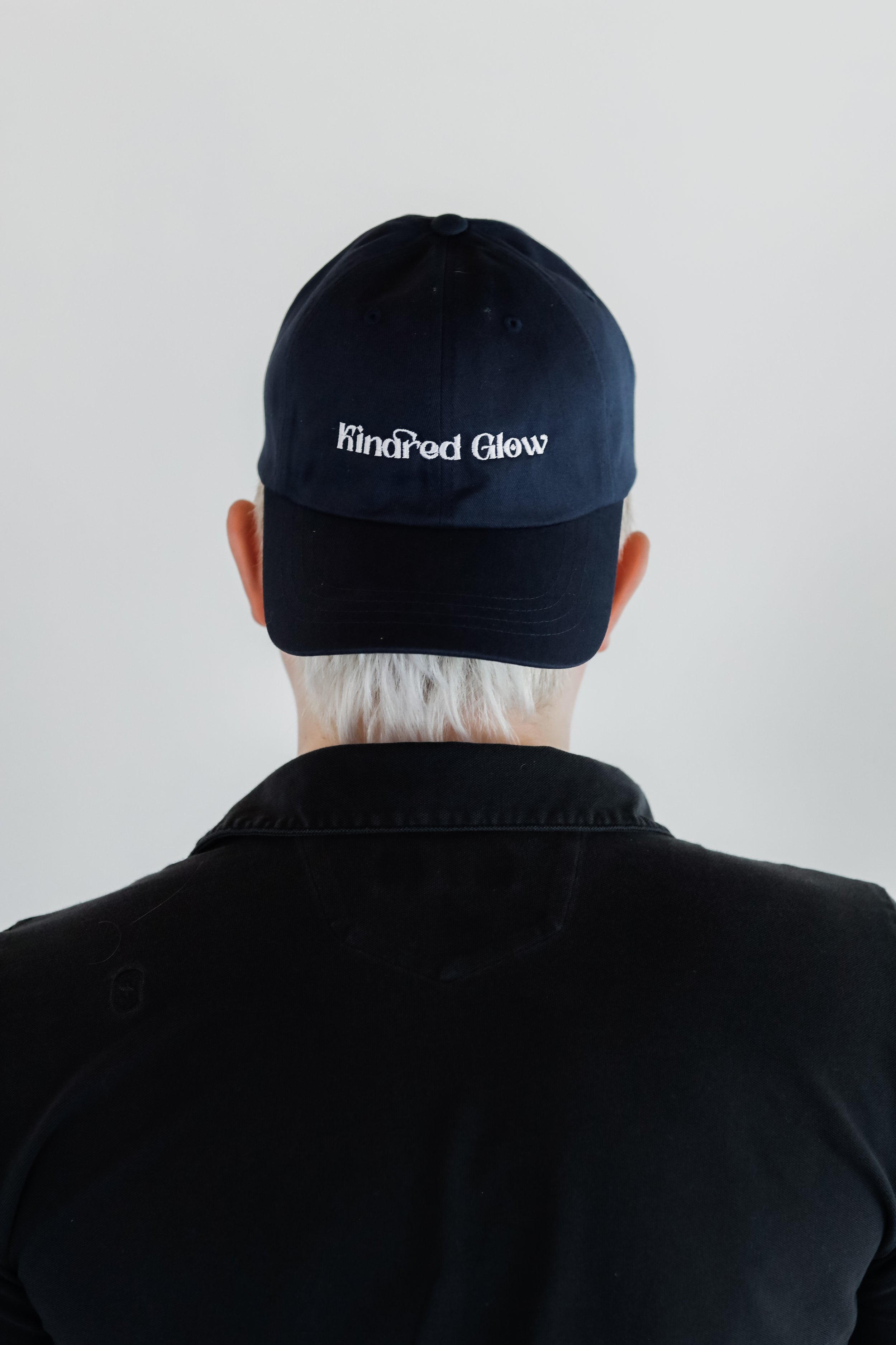 Person wearing a black baseball cap with white embroidered text 'kindred Glow' seen from behind, against a plain white wall, with short white hair visible