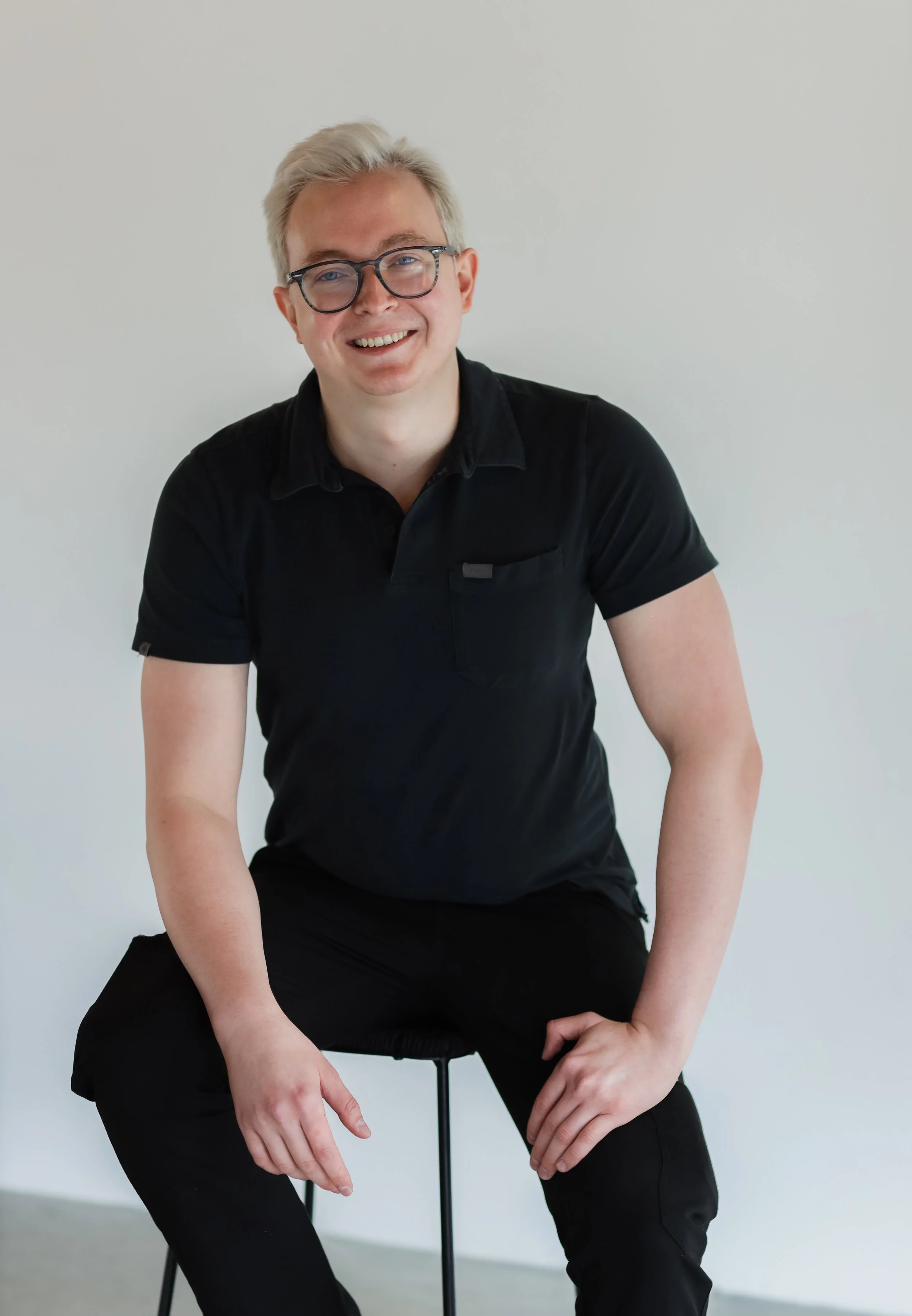 A smiling young man with blonde hair and glasses, wearing a black shirt, sitting on a stool against a plain white wall.