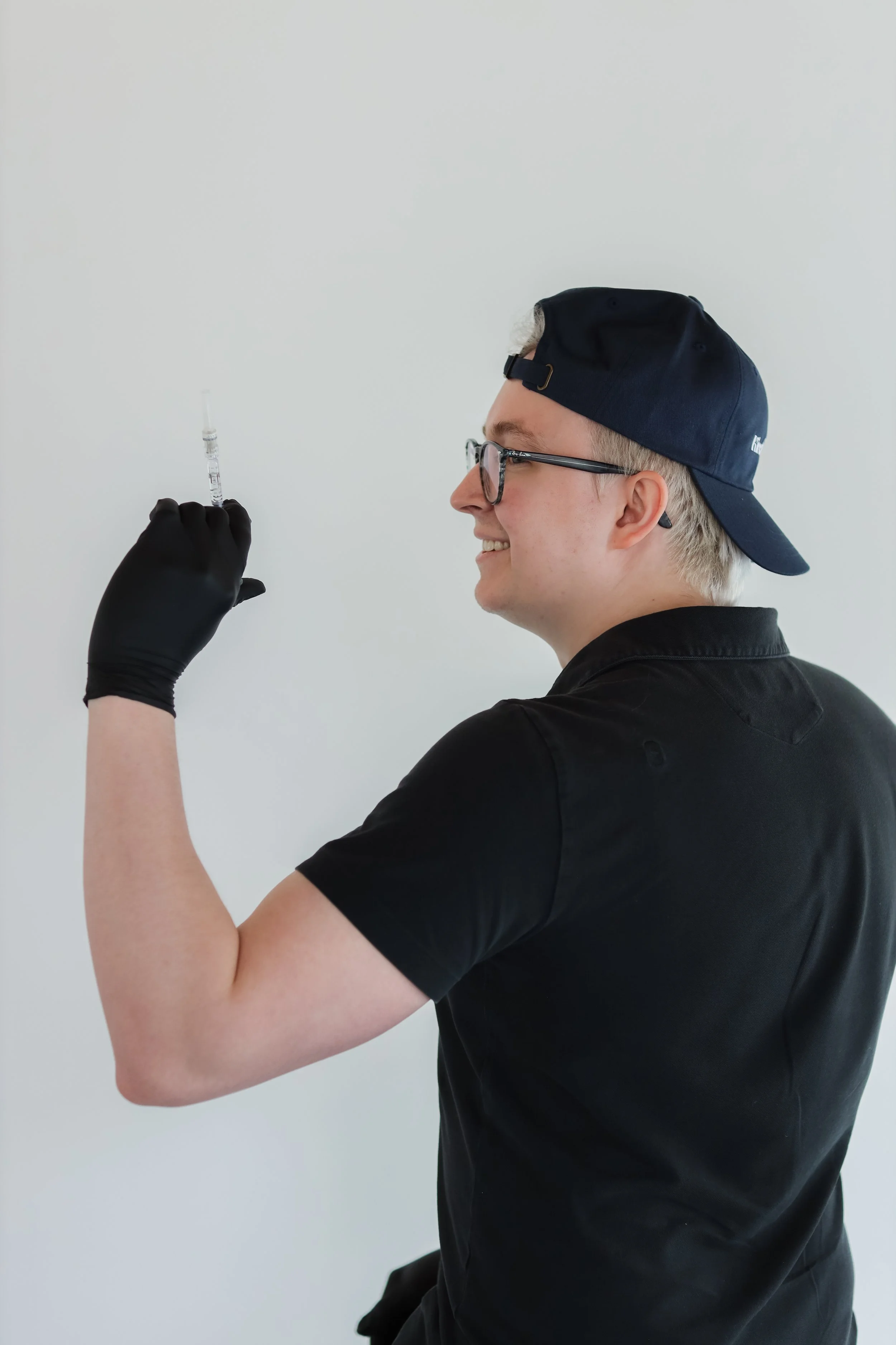 A young man wearing glasses, a black shirt, a backwards navy cap, and black gloves, smiling as he holds a syringe up in a white room.