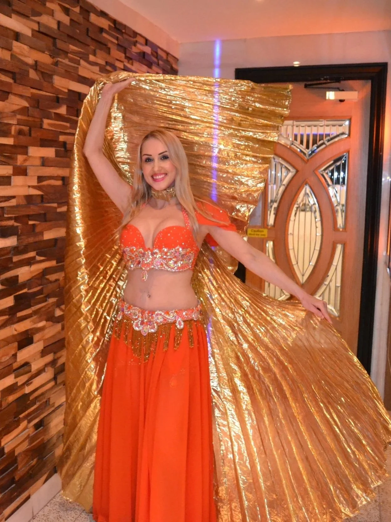 A woman in an orange costume with gold accents, holding a large gold fan, standing inside a building with a textured wooden wall and an ornate door.
