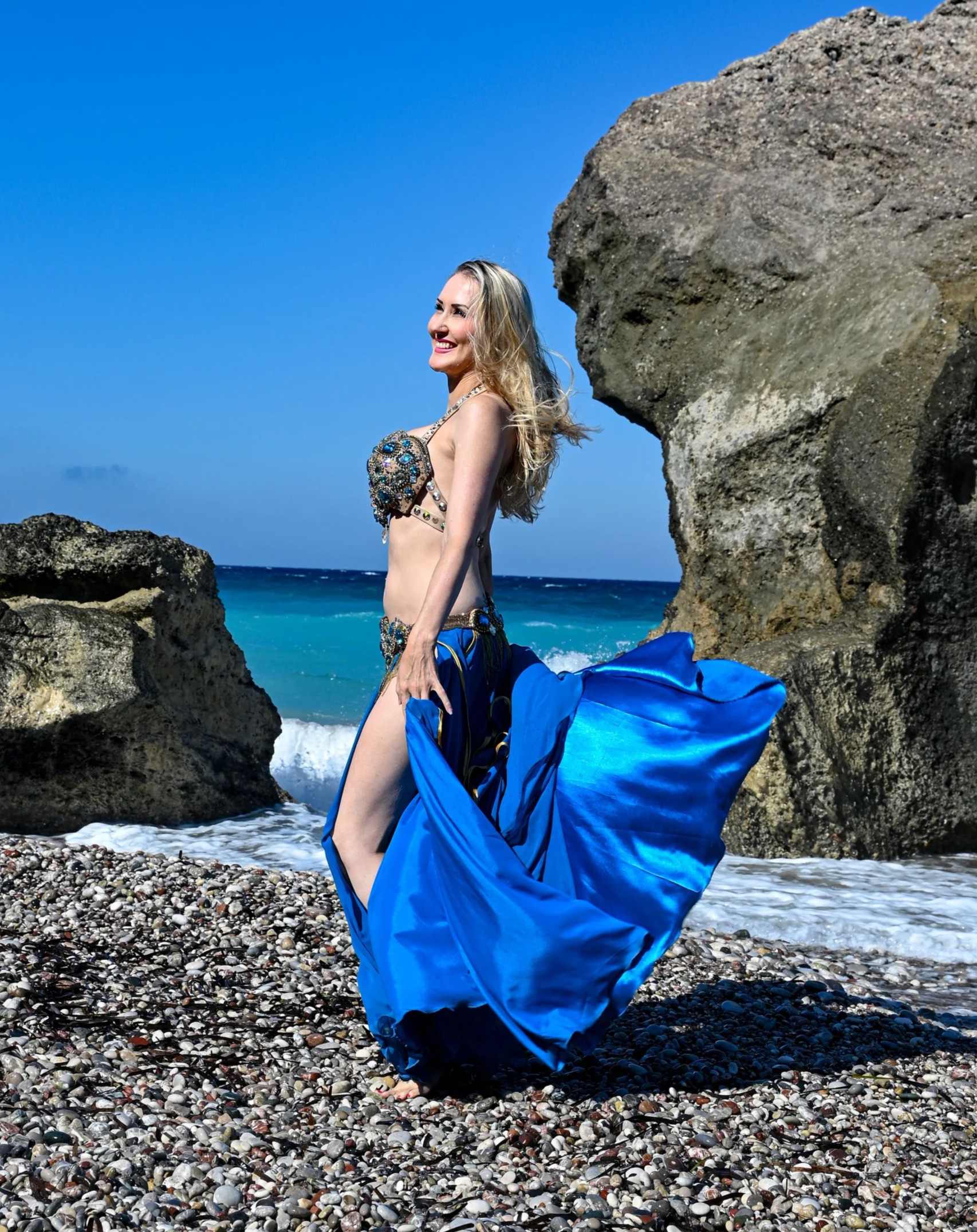 Woman in a blue skirt and bikini top standing on a pebble beach with rocks and the ocean in the background.