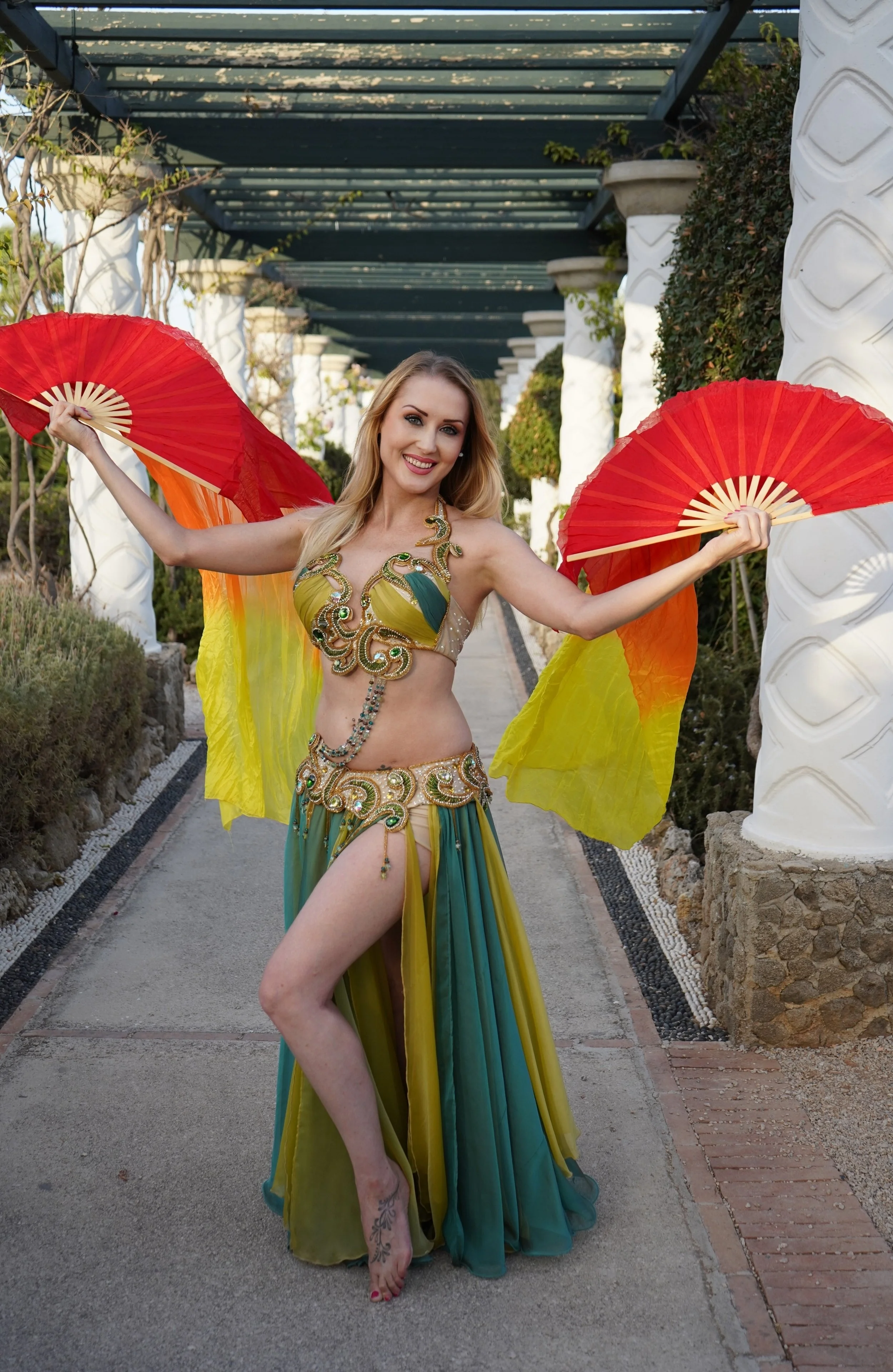 A woman in a colorful, elaborate belly dance costume holds red fans in each hand and smiles at the camera. She is standing in a garden walkway with white pillars and greenery around.