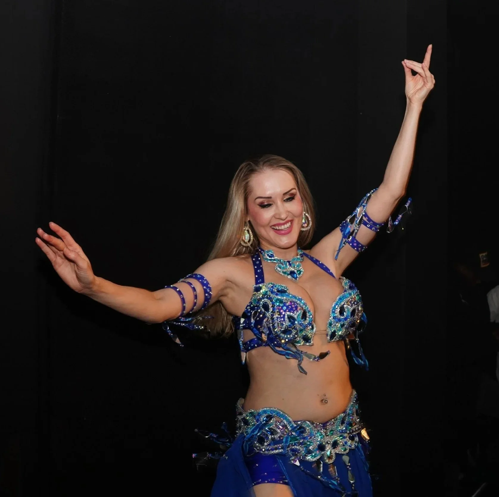 A woman wearing a blue, jeweled belly dance costume with jewelry, smiling and dancing against a black background.