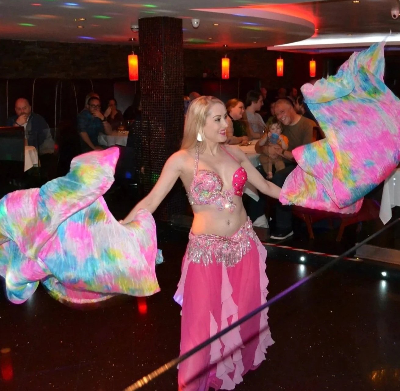 A belly dancer performing at a restaurant with an audience watching and dining. The dancer is wearing a pink costume with colorful decorations and holding large, colorful silk veils.