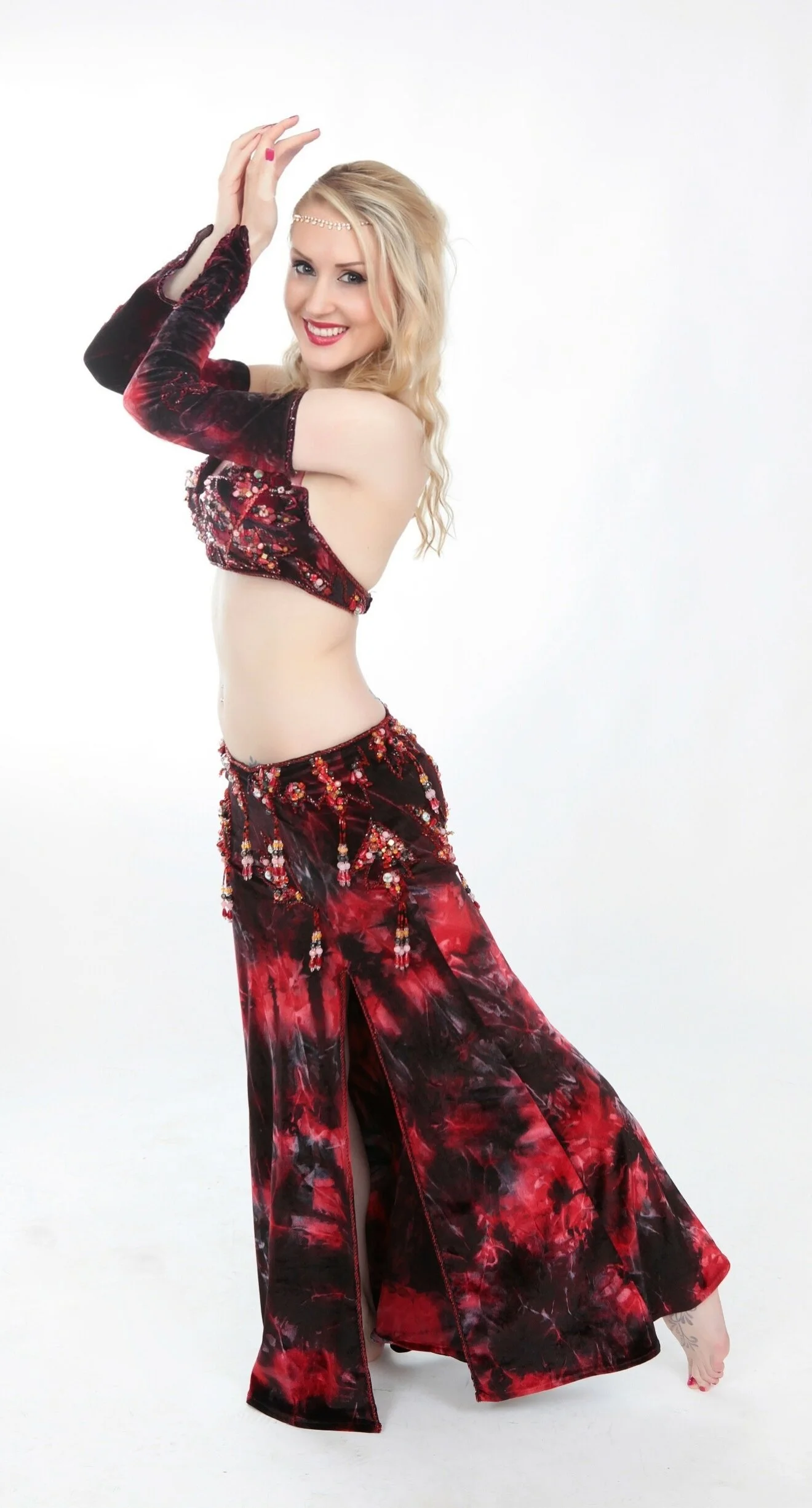 A woman with long blonde hair in a belly dance costume, wearing a red and black tie-dye top and matching skirt with beaded embellishments, posing with her hands above her head against a plain white background.