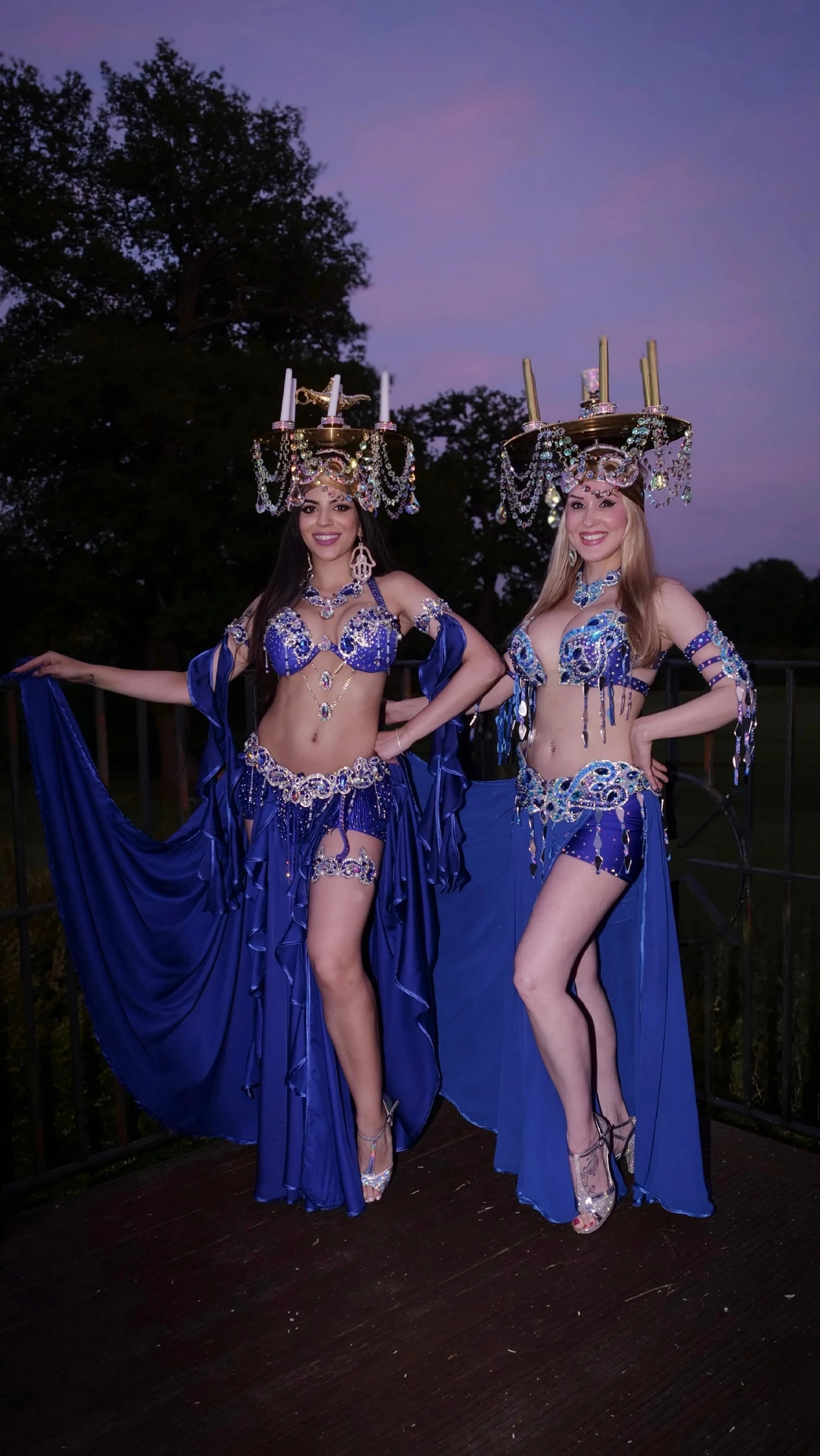 Two women in elaborate blue costumes with jewelry and large headpieces, standing outdoors during dusk, smiling at the camera.