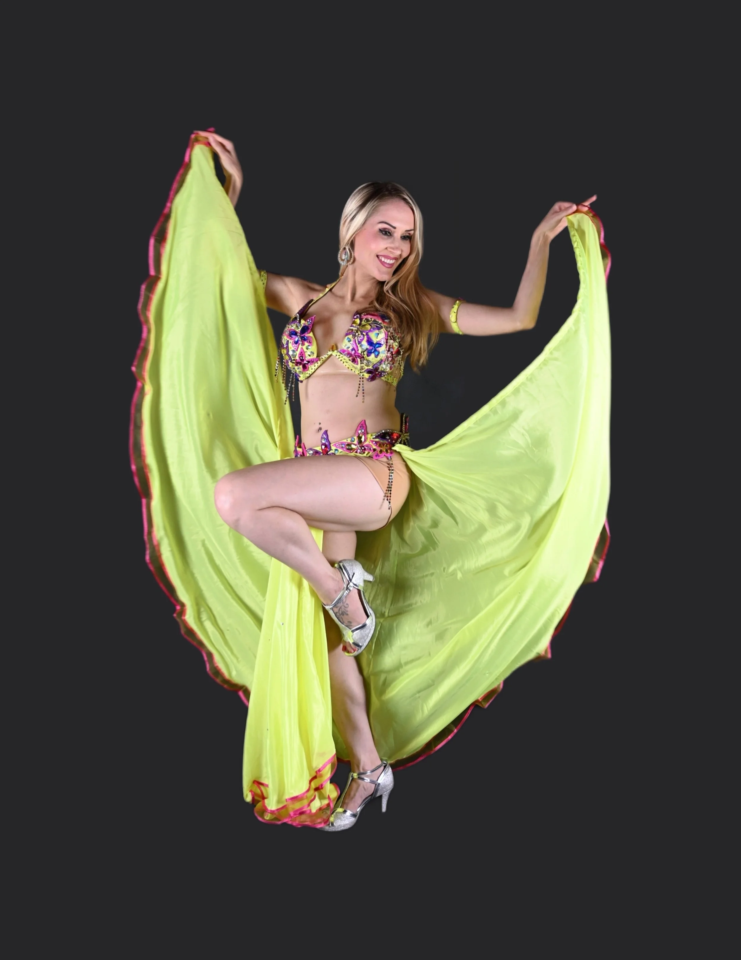 A woman dancing in a colorful costume, holding up a yellow flowing skirt, standing on one leg with a gray high heel shoe, smiling against a dark background.