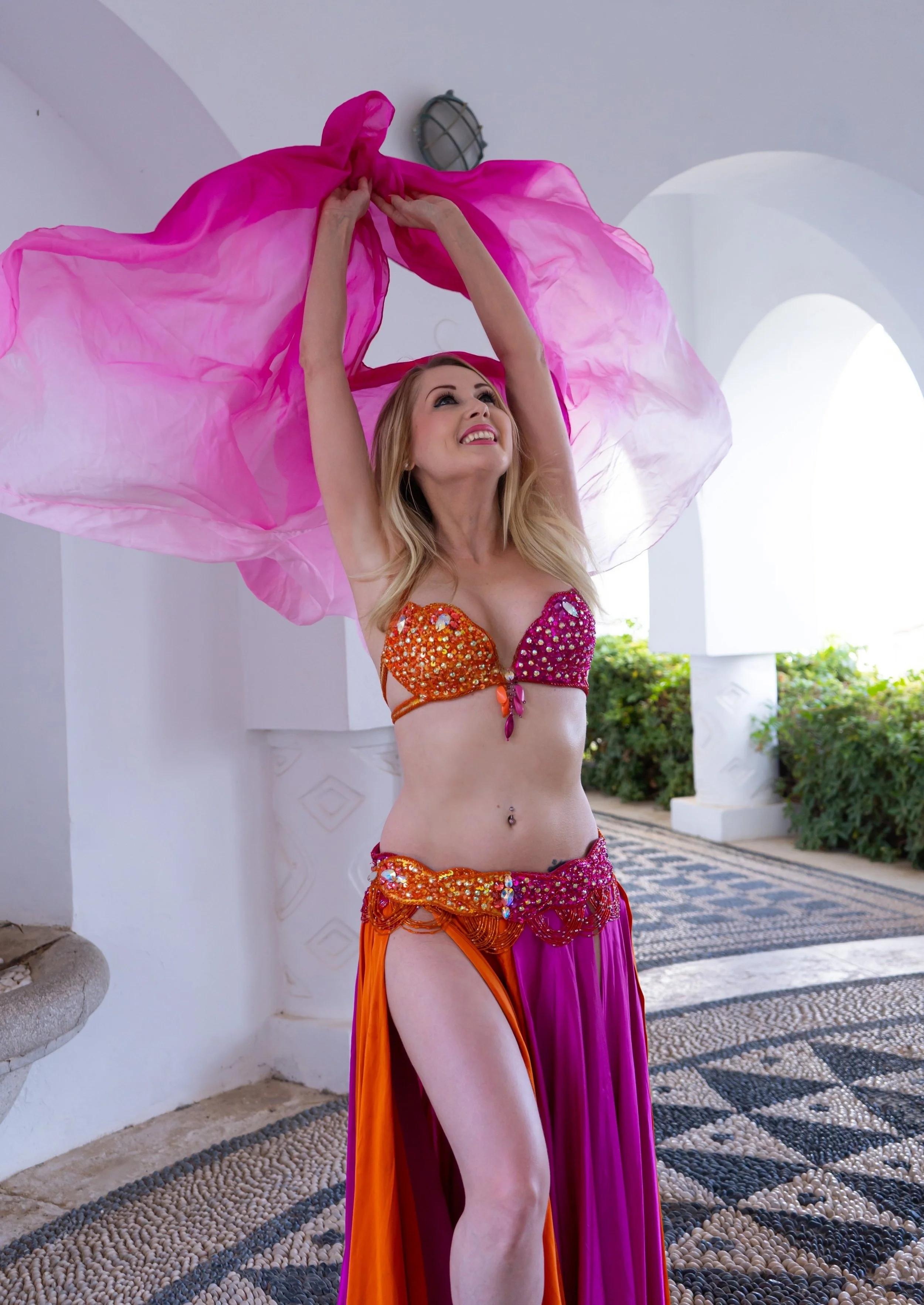A woman in colorful belly dance costume, smiling and holding a pink veil above her head.