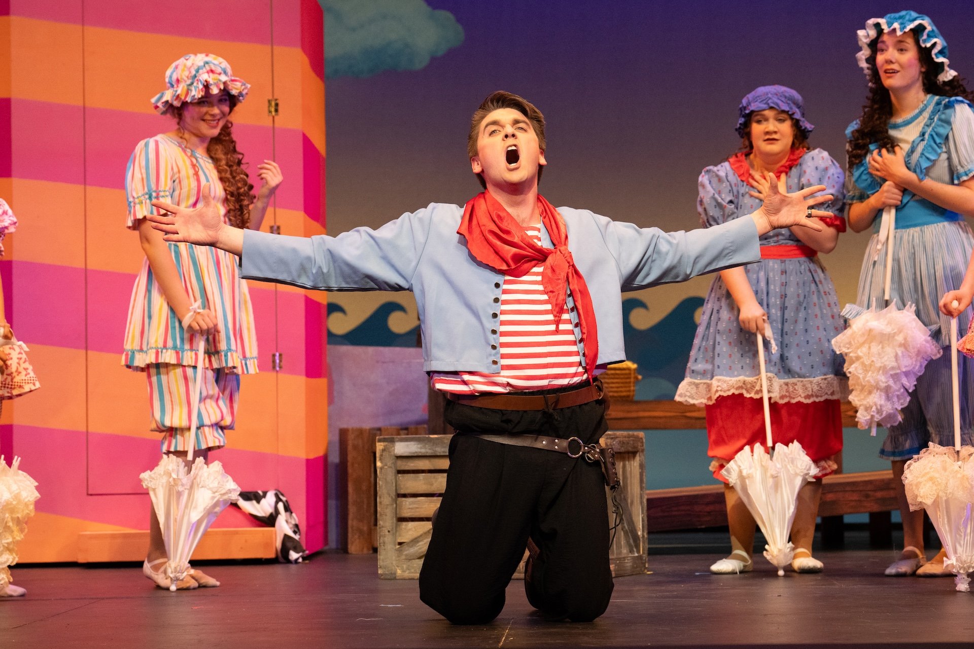 A theatrical stage scene featuring a man, possibly an actor, kneeling with arms outstretched and mouth open, surrounded by women dressed in colorful, old-fashioned dresses and bonnets, indicating a performance likely set in a whimsical or historical context.