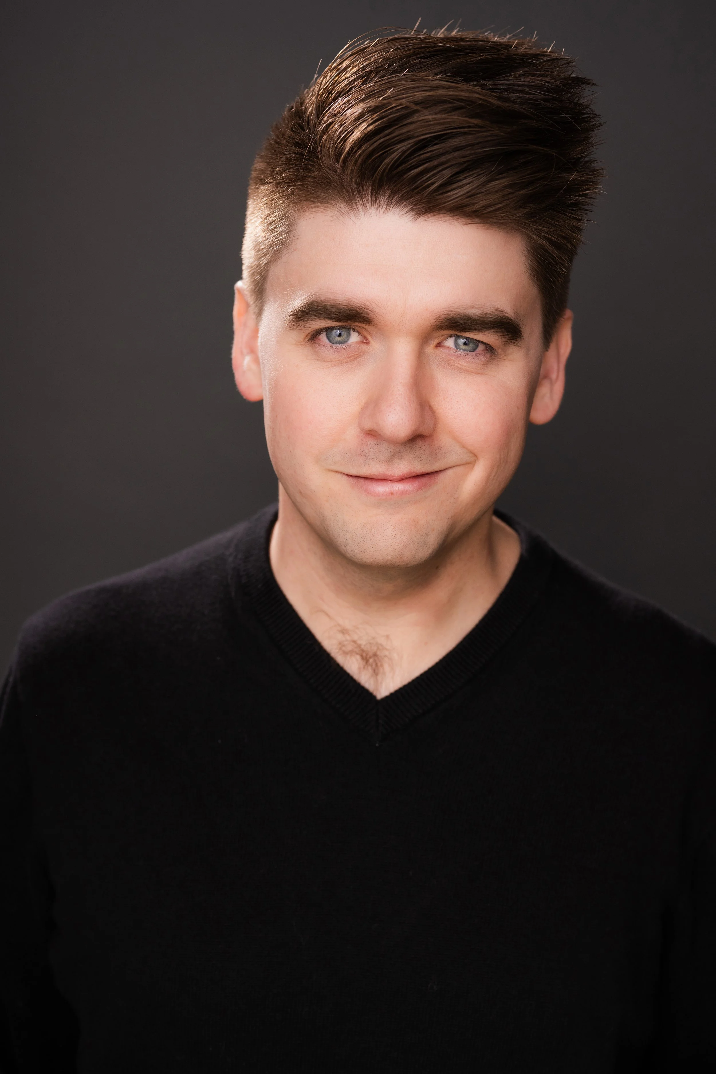 A young man with light skin, dark styled hair, and blue eyes, wearing a black V-neck sweater, smiling slightly against a dark background.