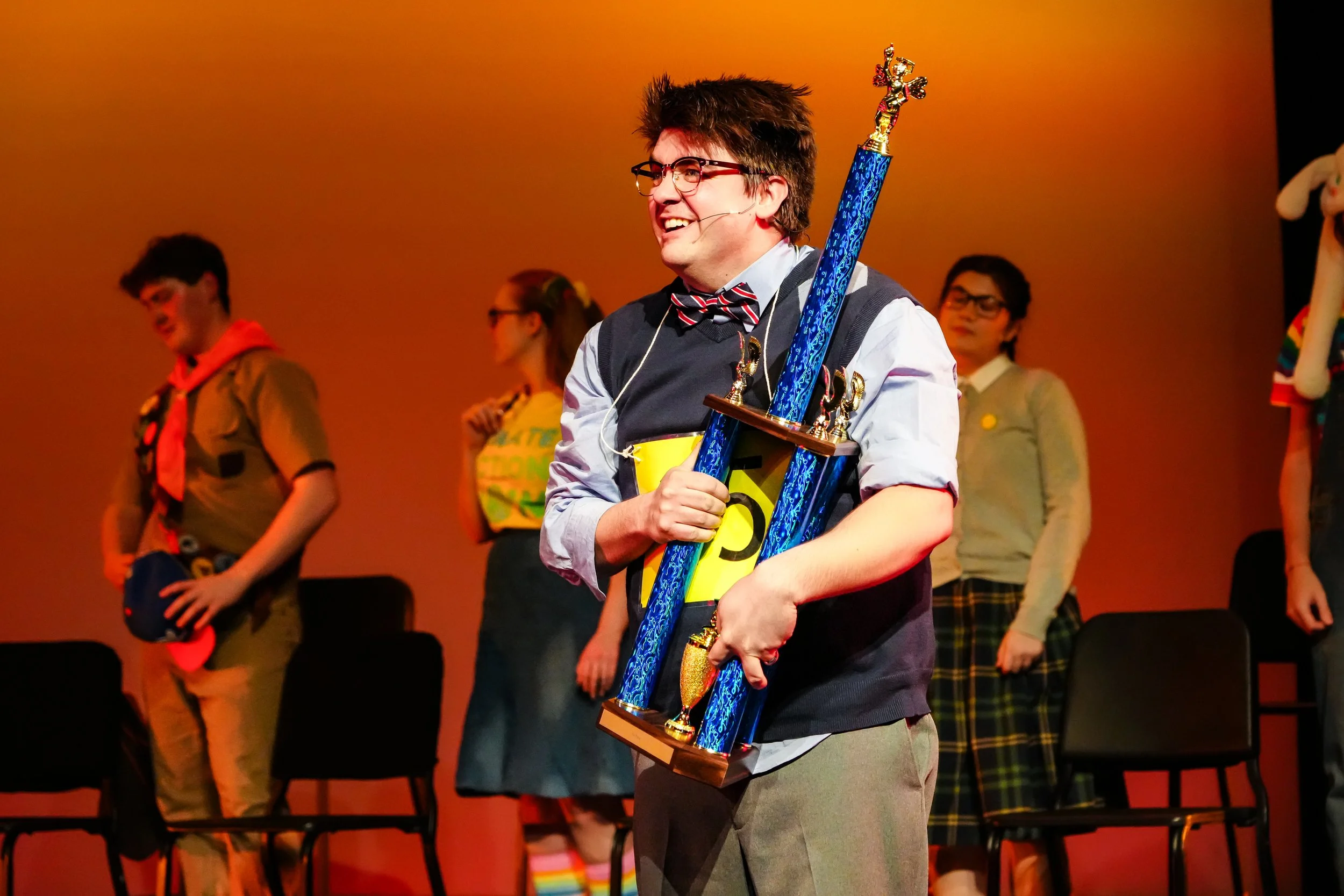 Young man with glasses, wearing a bow tie and a headset microphone, holding a large trophy with a blue column and a gold statue on top, on stage with other people in the background.