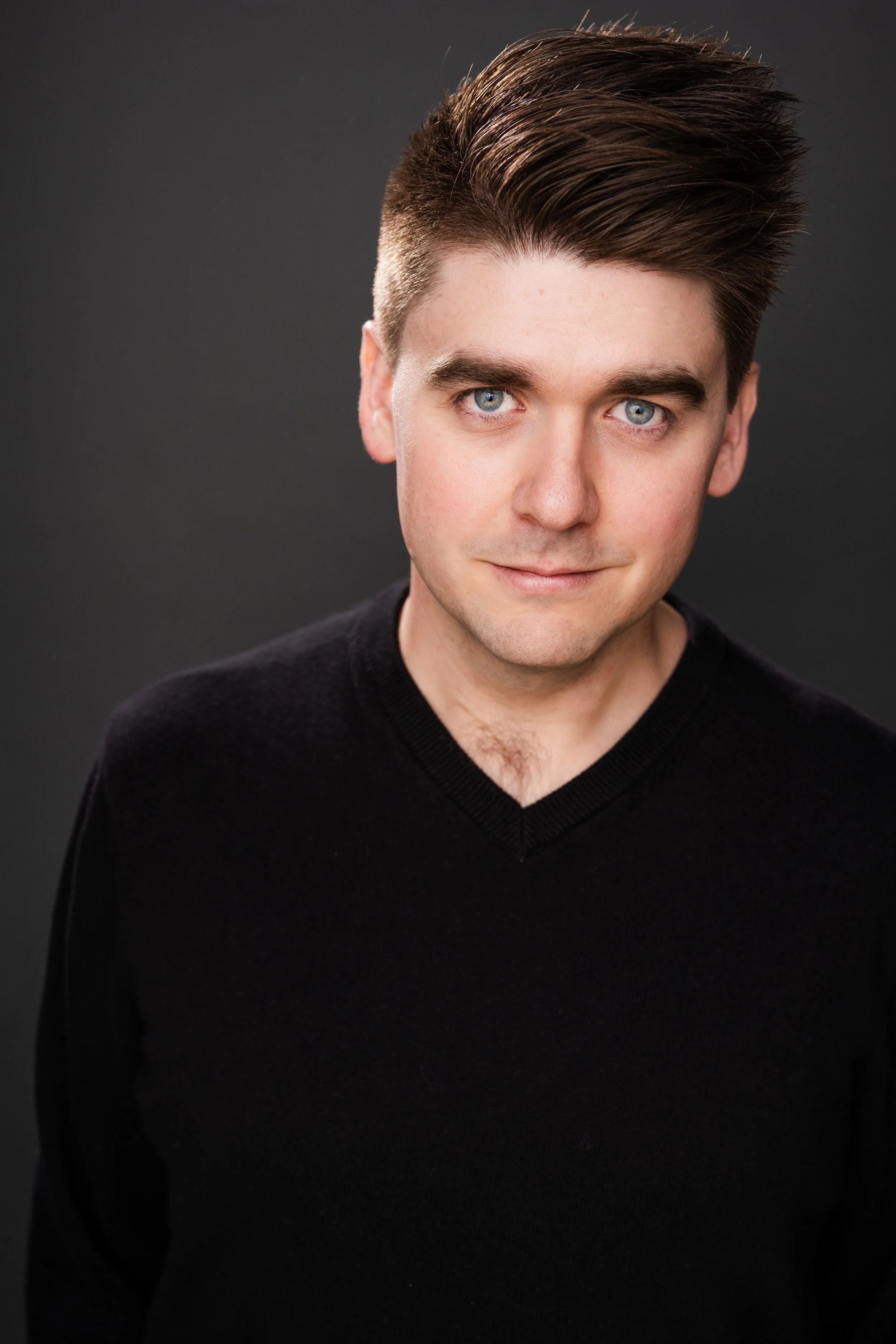 A young man with styled brown hair, blue eyes, and fair skin, wearing a black V-neck sweater, is gazing at the camera against a dark background.