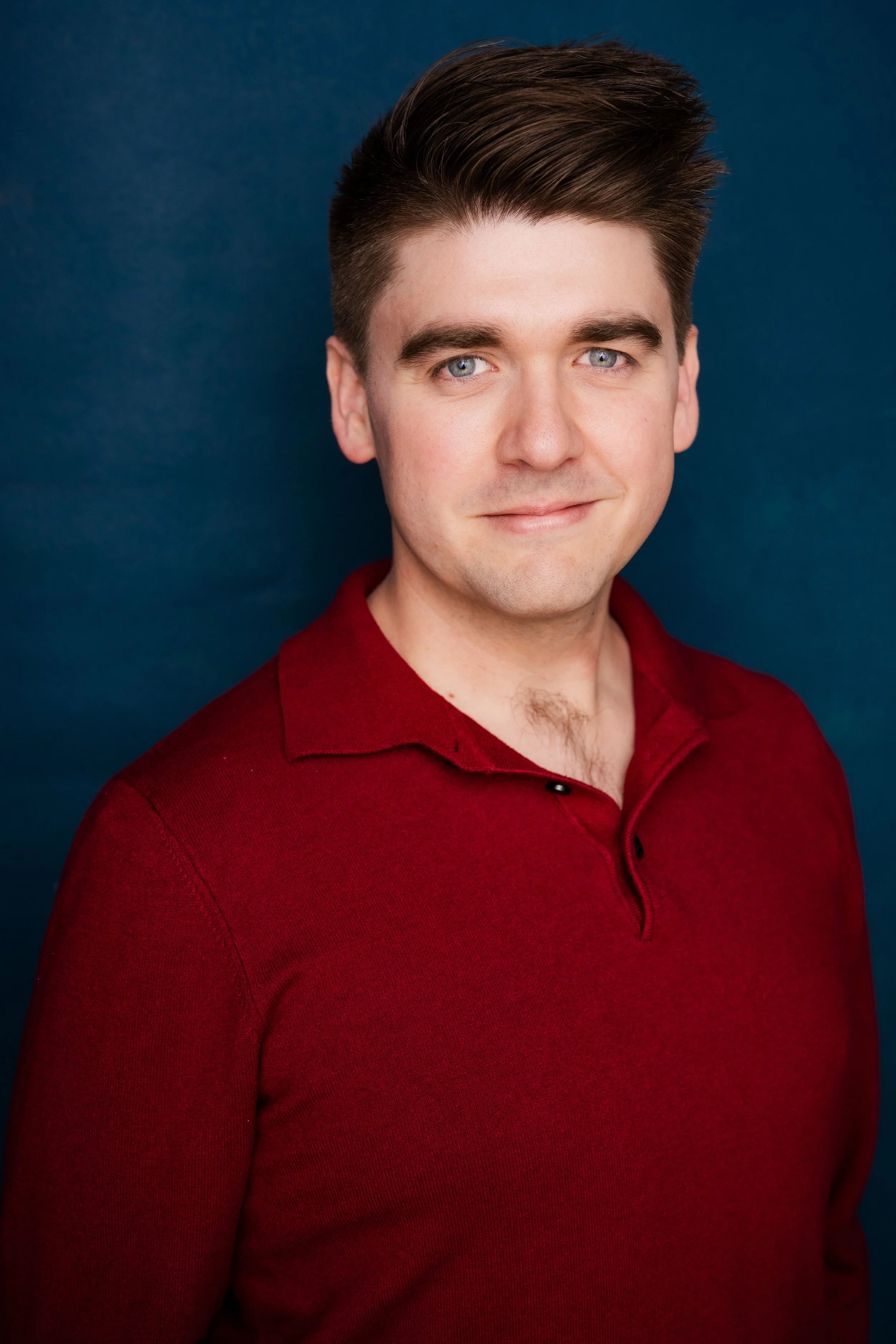 A young man with short, styled brown hair and blue eyes, wearing a red polo shirt against a dark blue background.