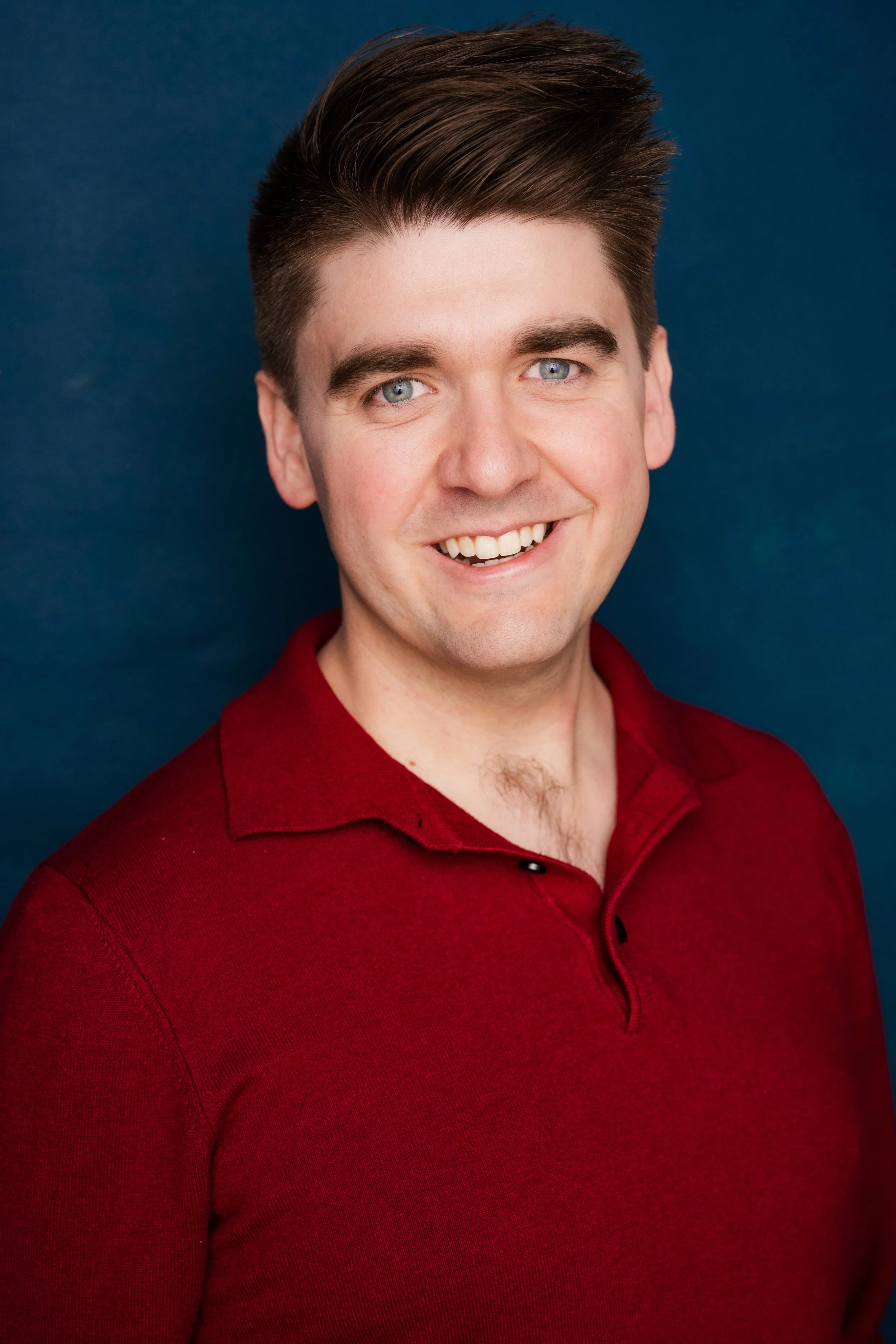 A young man with blue eyes, dark brown hair styled upwards, smiling, wearing a red collared shirt against a dark blue background.