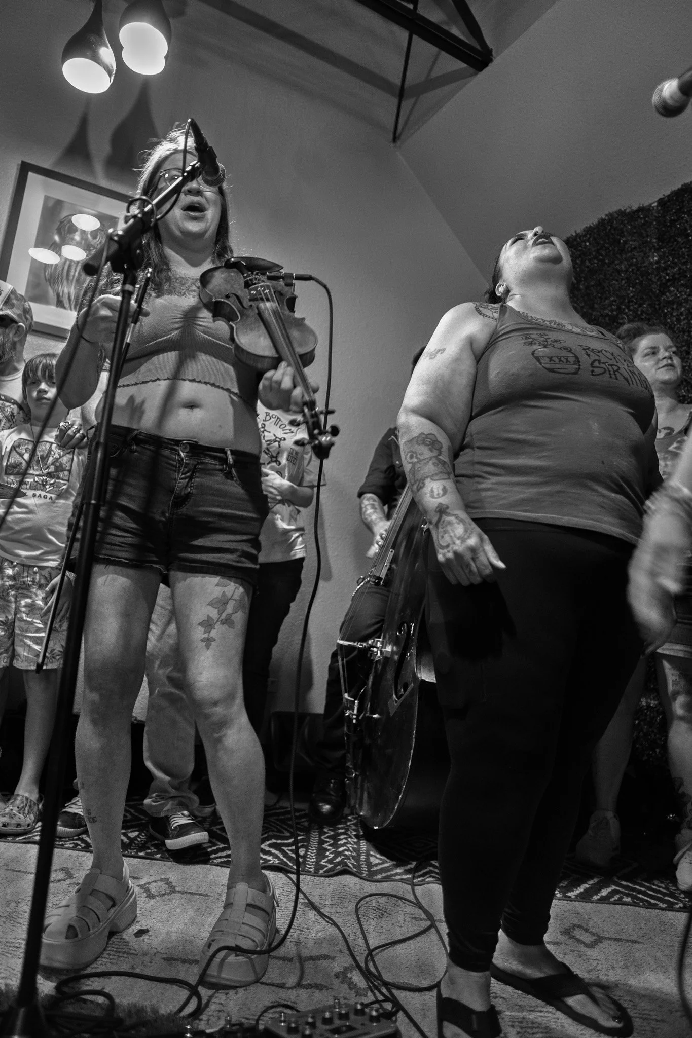 Black and white photo of two women performing music, one playing a violin and the other singing with head tilted back, in a room with a patterned rug and pendant lights overhead.