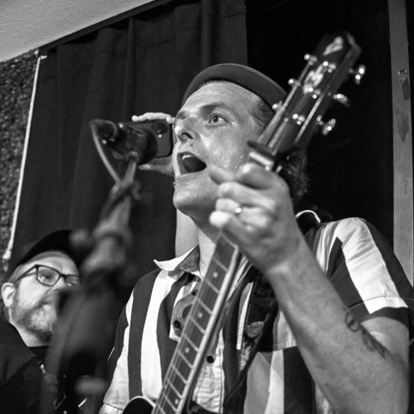 A musician singing into a microphone while playing an acoustic guitar, with a man with glasses in the background.