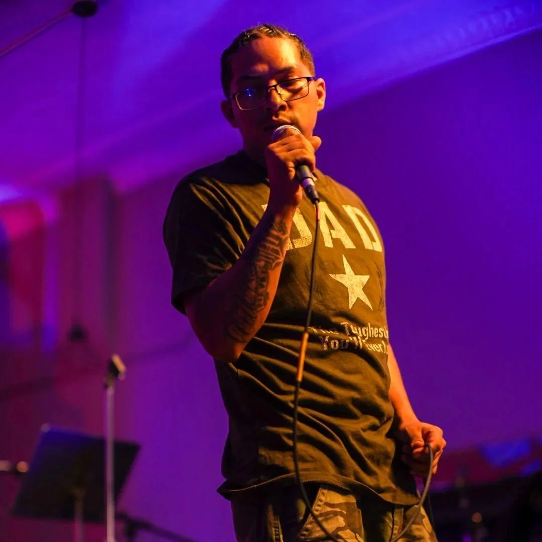 Person with glasses holding a microphone on stage, singing or speaking, with purple and blue stage lighting.
